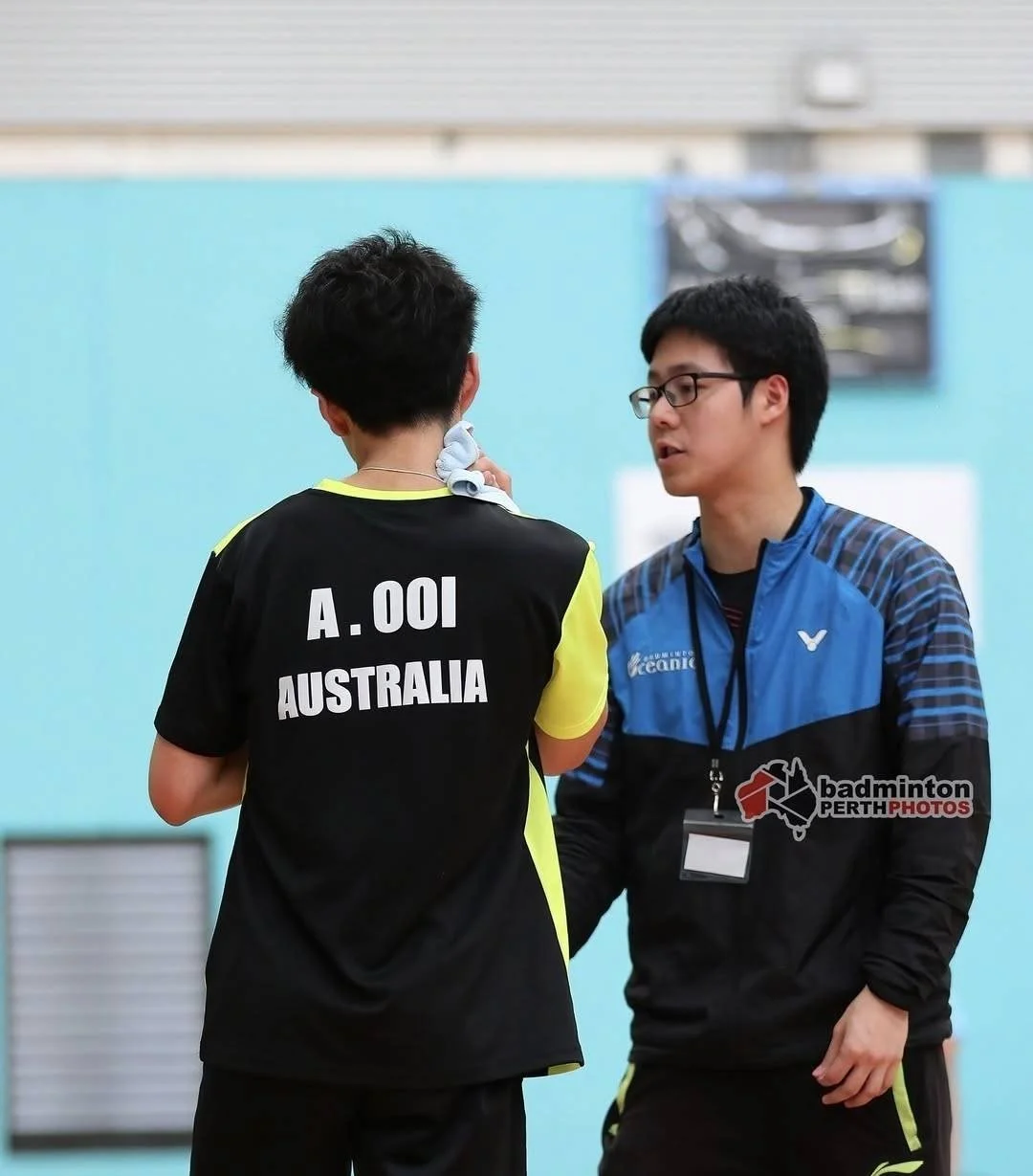 A tennis player wearing a black and yellow uniform with 'A. Ooi Australia' on the back is being attended to by a coach or official wearing glasses and a blue jacket in a sports hall.