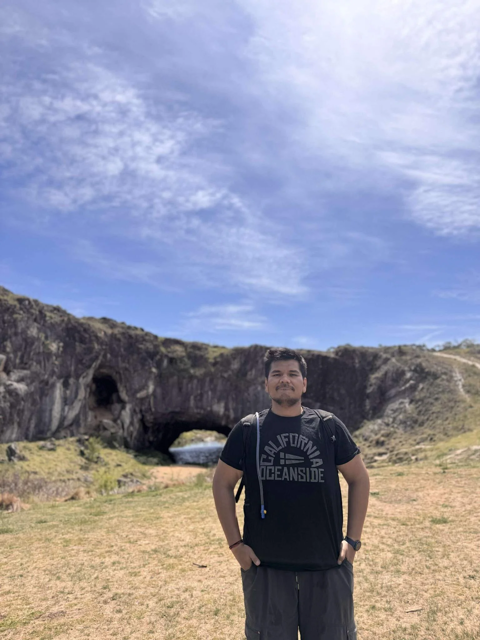 A man standing outdoors in front of a natural rock formation with a small ocean cave and arch in the background, under a blue sky with scattered clouds.