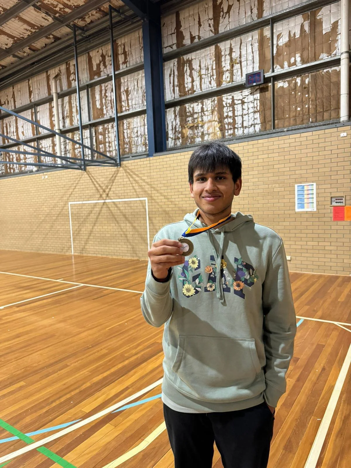 A young man holding a gold medal stands in a gymnasium with a wooden floor and brick wall.