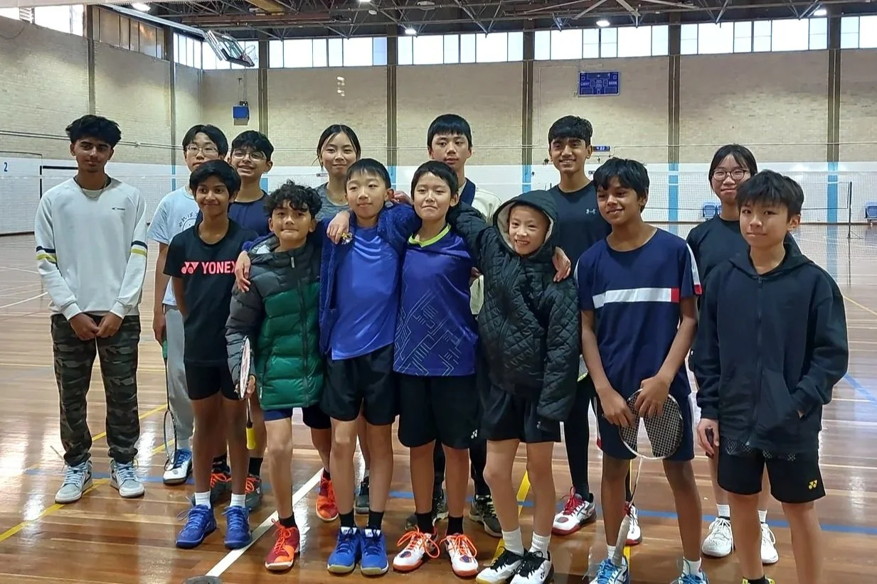 Group of young boys and girls standing together on an indoor badminton court, smiling at the camera.
