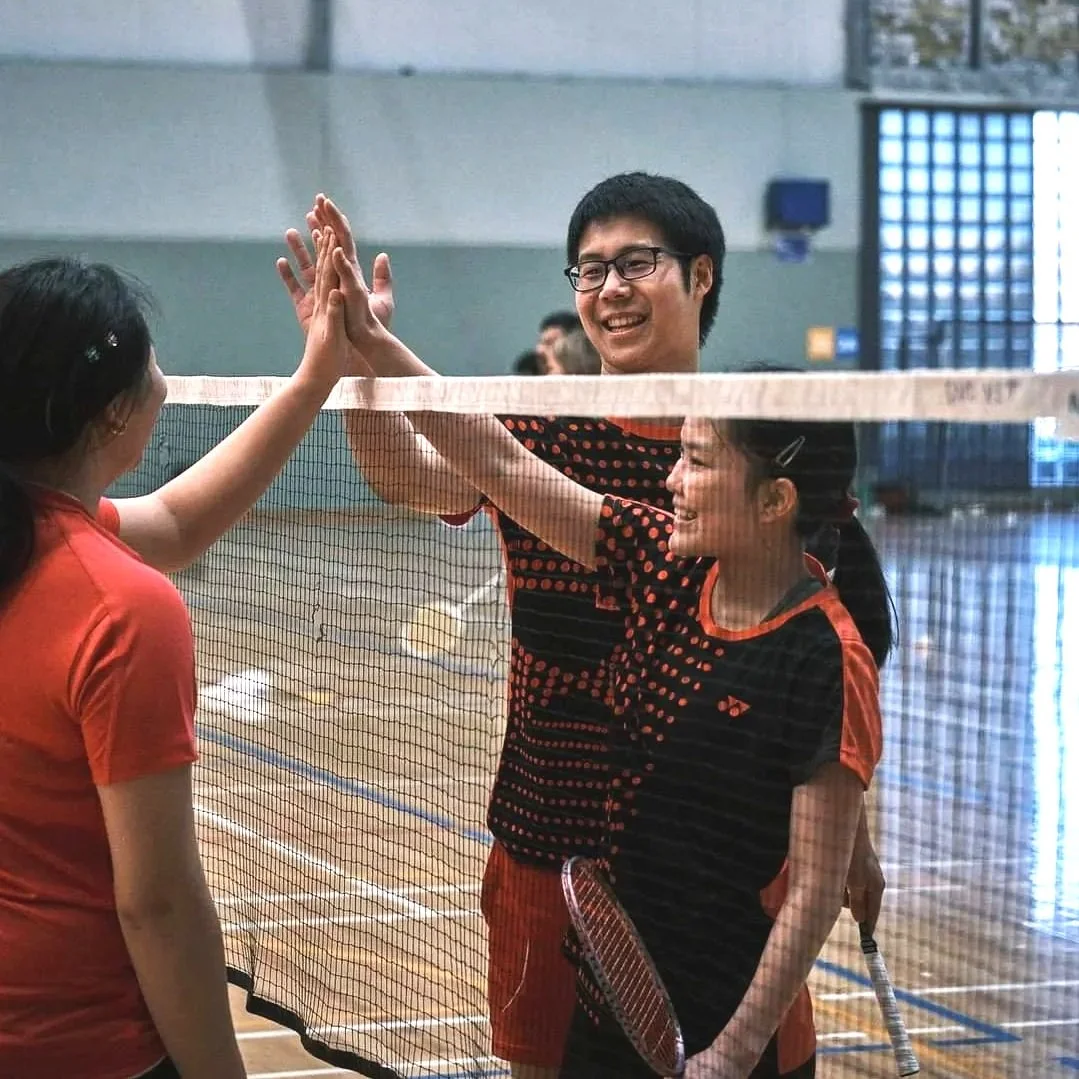 Three people in a gymnasium high-fiving on a badminton court, two women and one man, all smiling and holding badminton rackets.