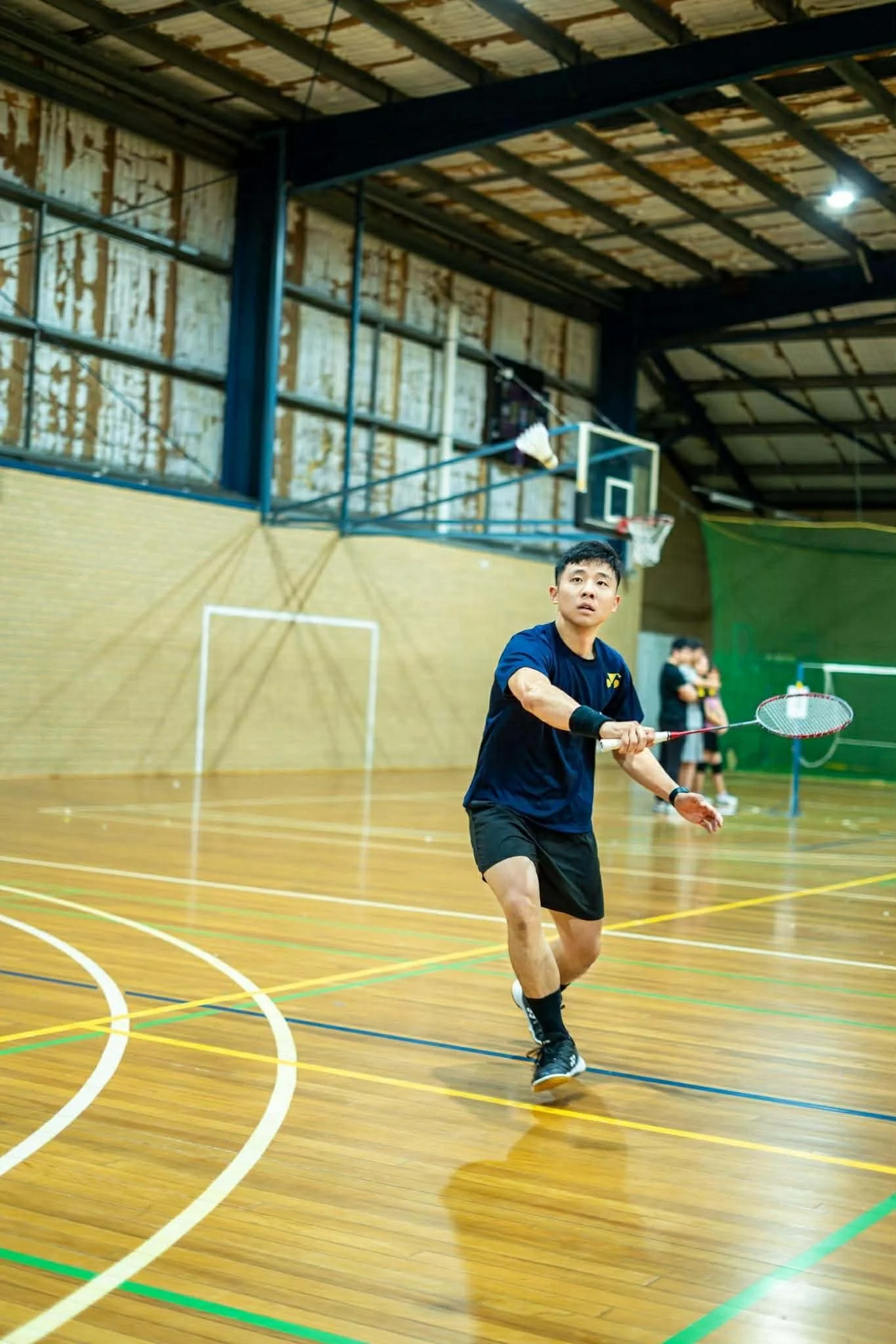 A young man in athletic clothing playing badminton indoors, holding a badminton racket, with a basketball hoop and two other people in the background on a wooden court.