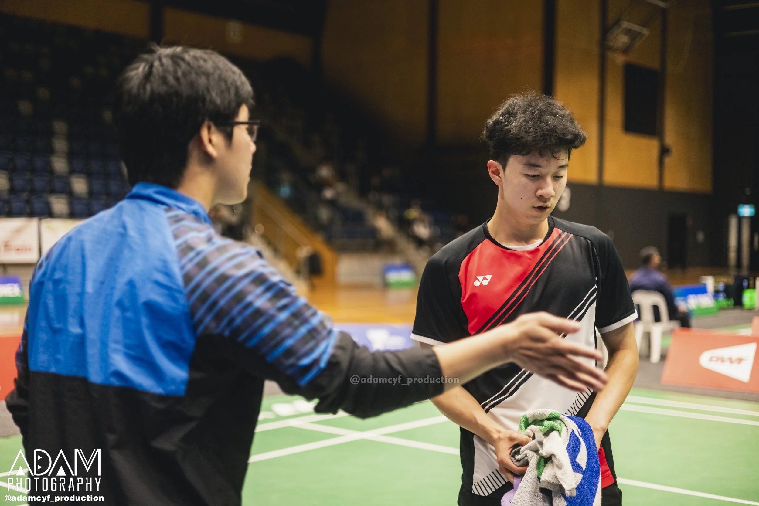 Two young men in a badminton court, one in a black and red sports shirt holding towels, talking while the other, wearing glasses and a blue jacket, gestures toward him. A gymnasium with seating and banners is in the background.