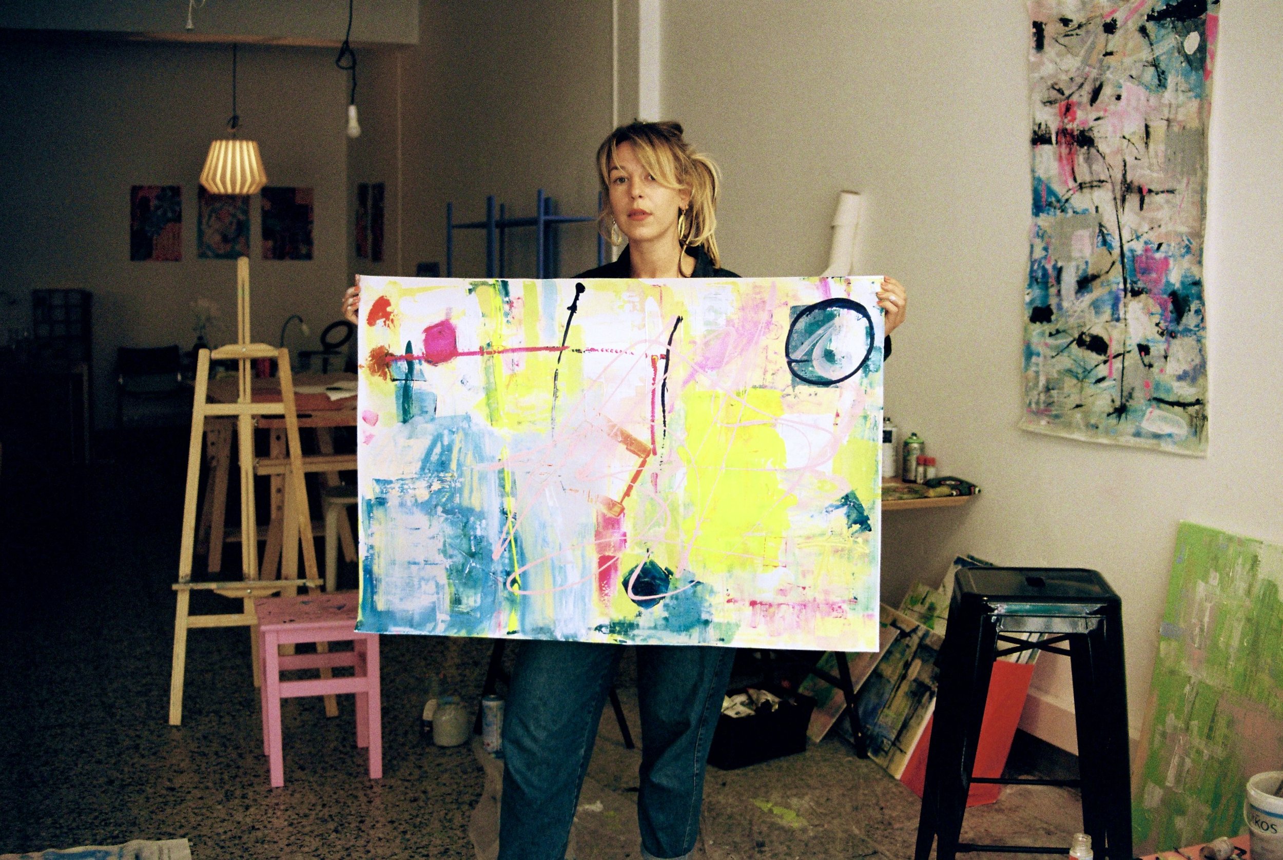 Künstler hält ein großes, buntes abstraktes Gemälde in einem Atelier. Im Hintergrund sind weitere Gemälde und Malmaterialien sichtbar.