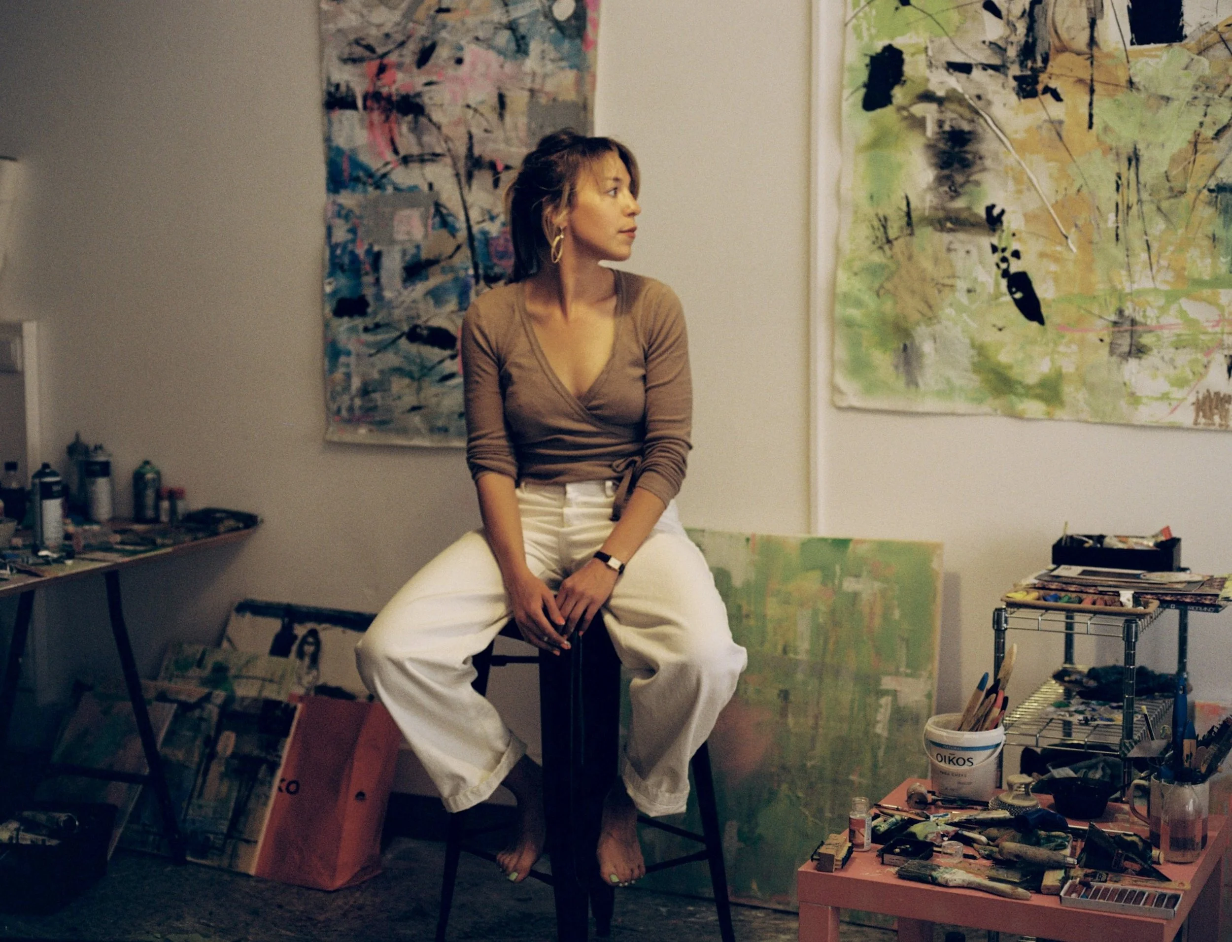 Eine Frau sitzt auf einem Stuhl in einem Kunststudio mit abstrakten Gemälden im Hintergrund, umgeben von Malutensilien und Leinwänden.