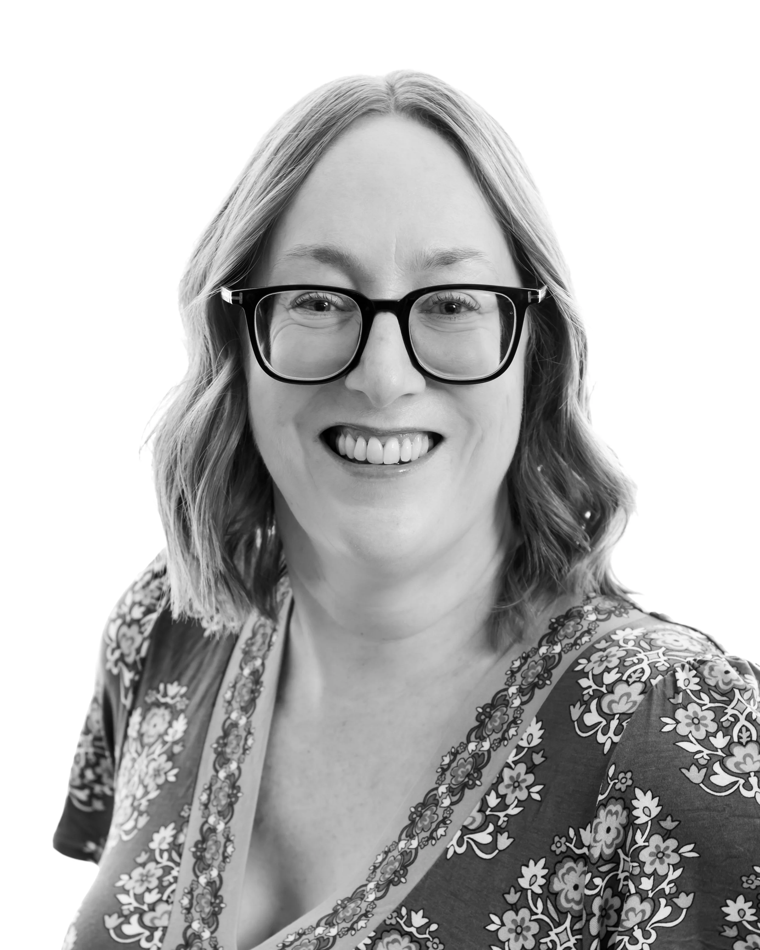 Black and white portrait of a woman with glasses, shoulder-length hair, smiling, wearing a floral patterned top.