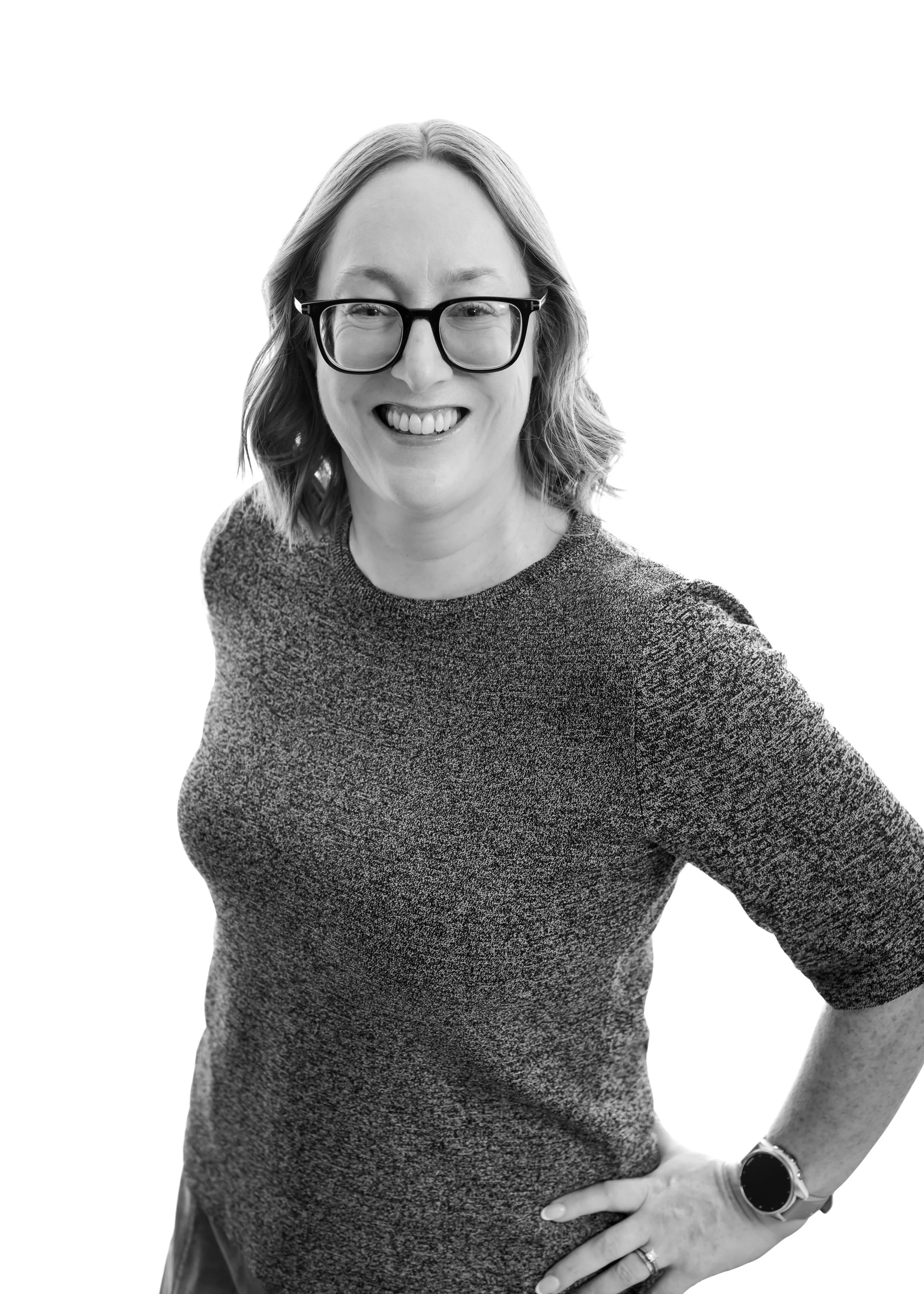 Black and white photo of a smiling woman wearing glasses, a patterned shirt, a wristwatch, and a ring, with her hand on her hip, looking directly at the camera.