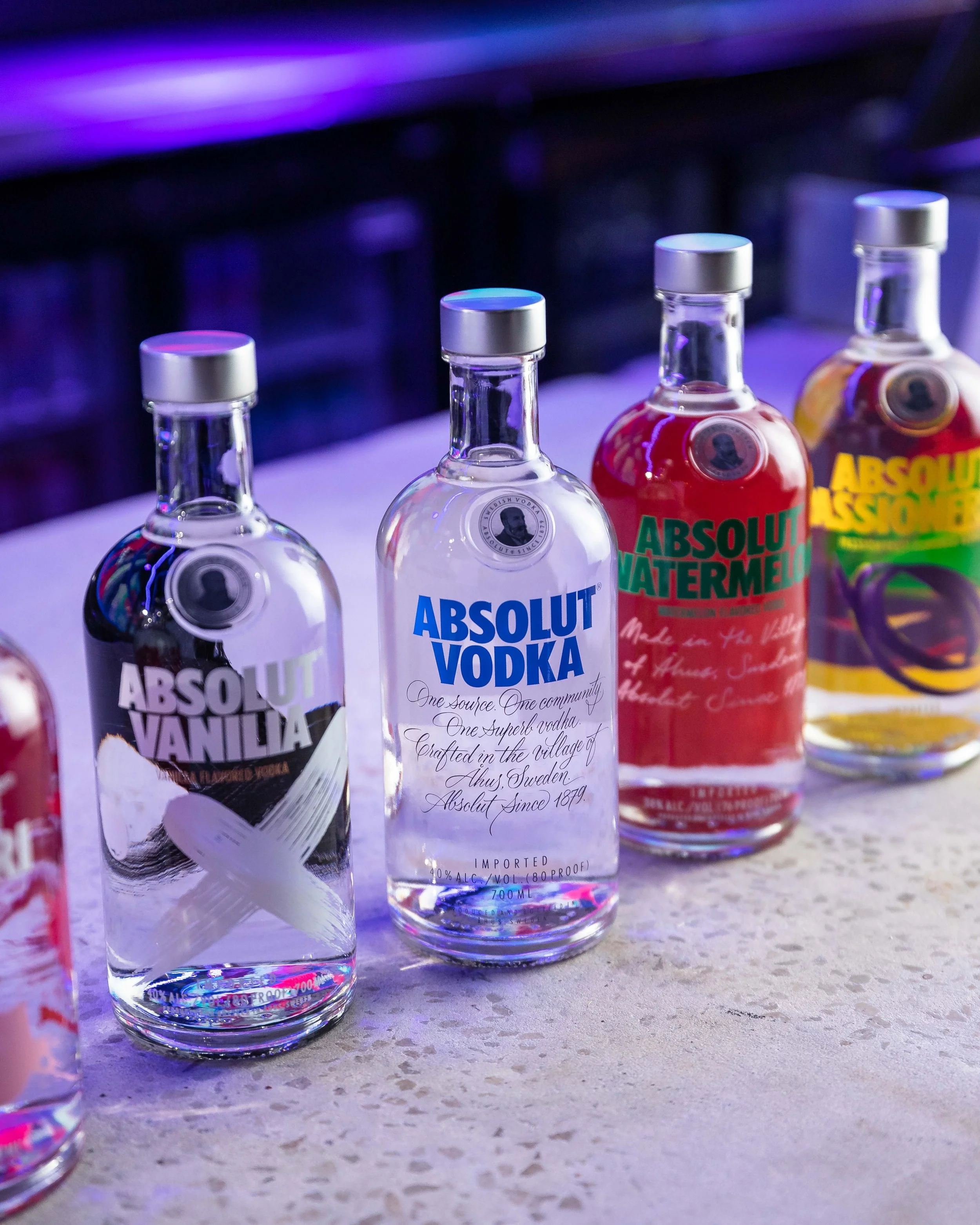 Four bottles of flavored vodka arranged on a counter with colorful lighting, including vanilla, vodka, watermelon, and Absolut Lemon flavors.
