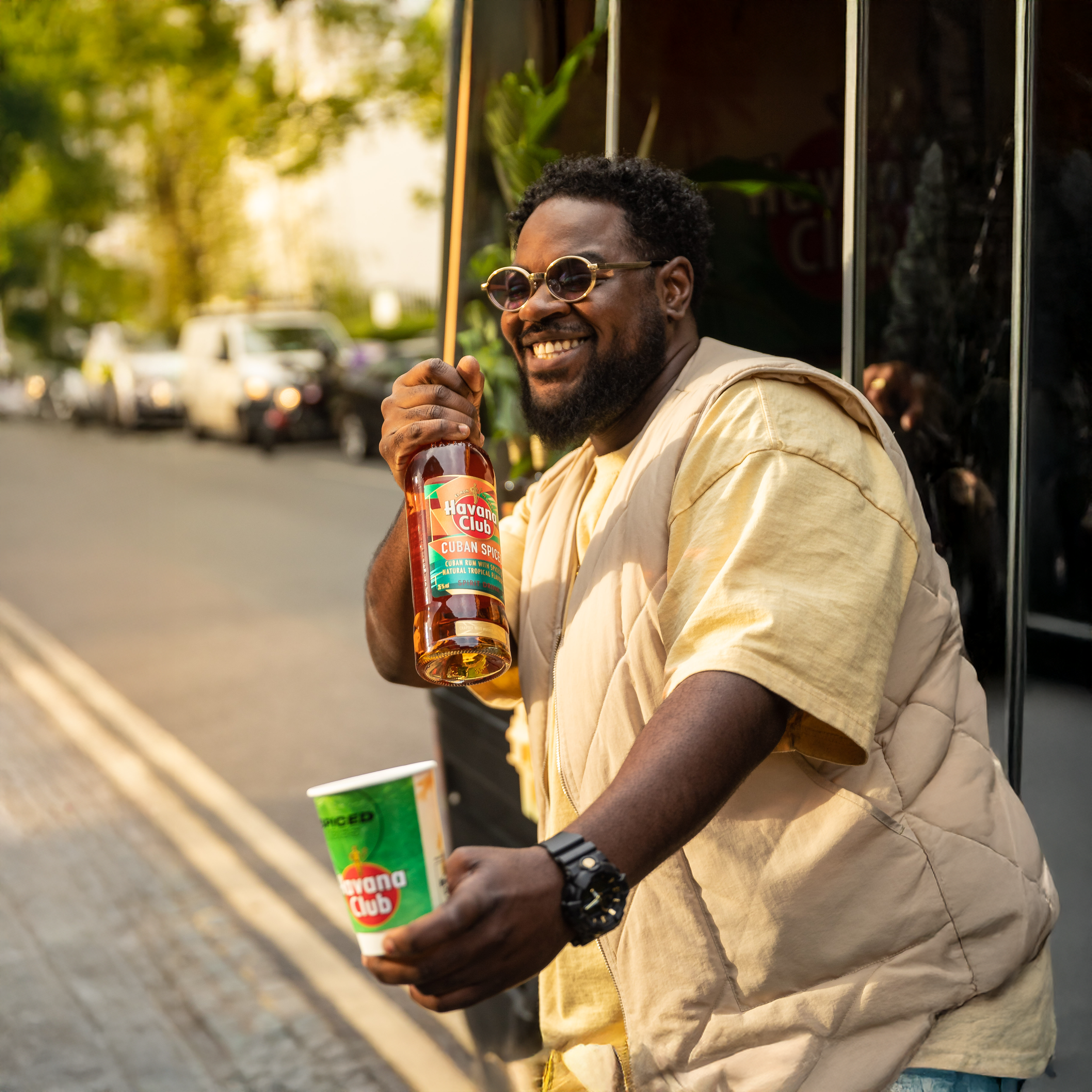 A man smiling and holding a bottle of Havana Club rum in one hand and a cup in the other, standing outside on a city street.
