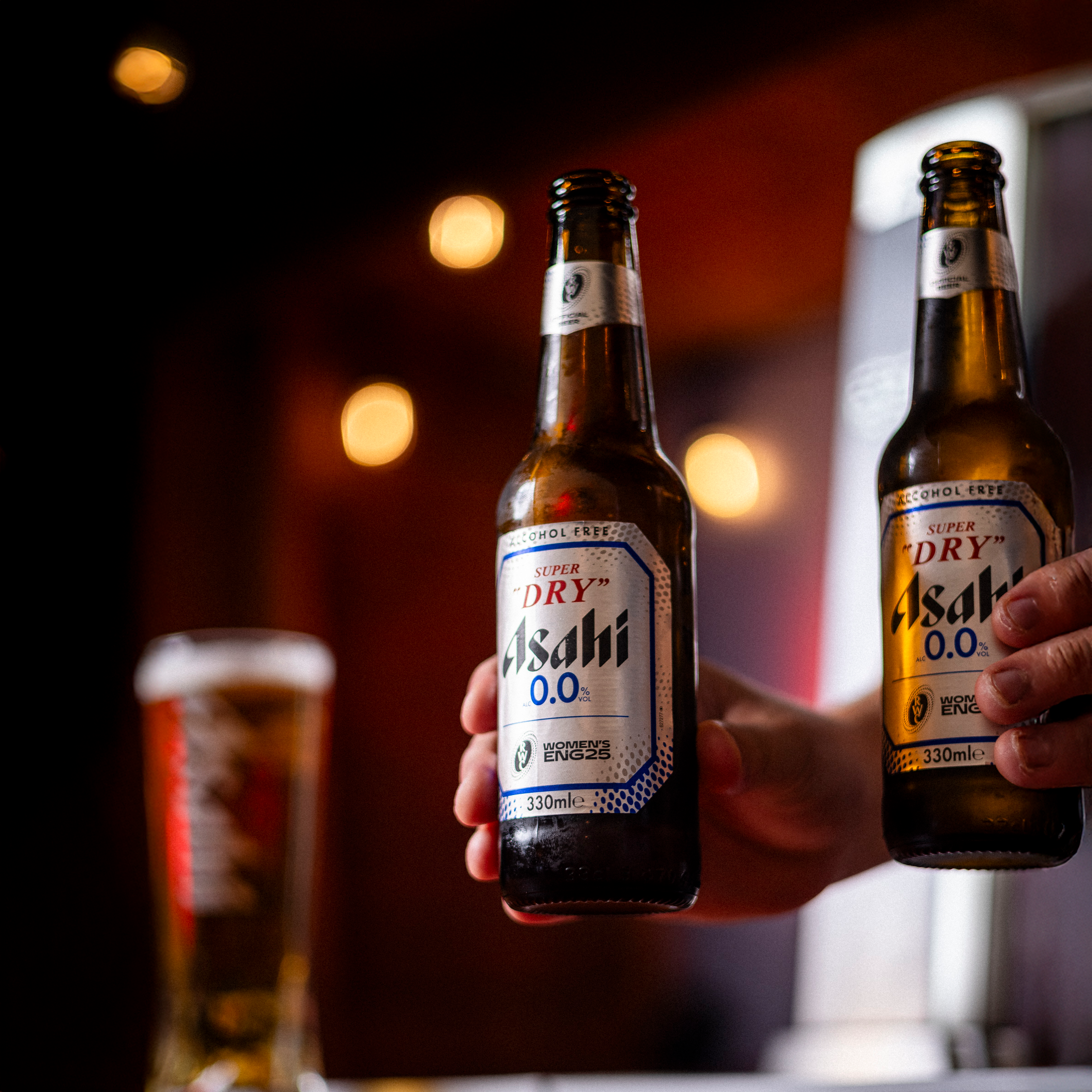 Two people holding bottles of Asahi alcohol-free beer with a blurred background of a glass of beer and warm lights.