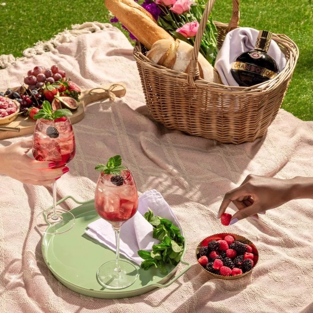 A picnic setup on a pink blanket with two glasses of berry drinks, a bowl of mixed berries, a basket with bread, flowers, and a bottle of Chambord liqueur.