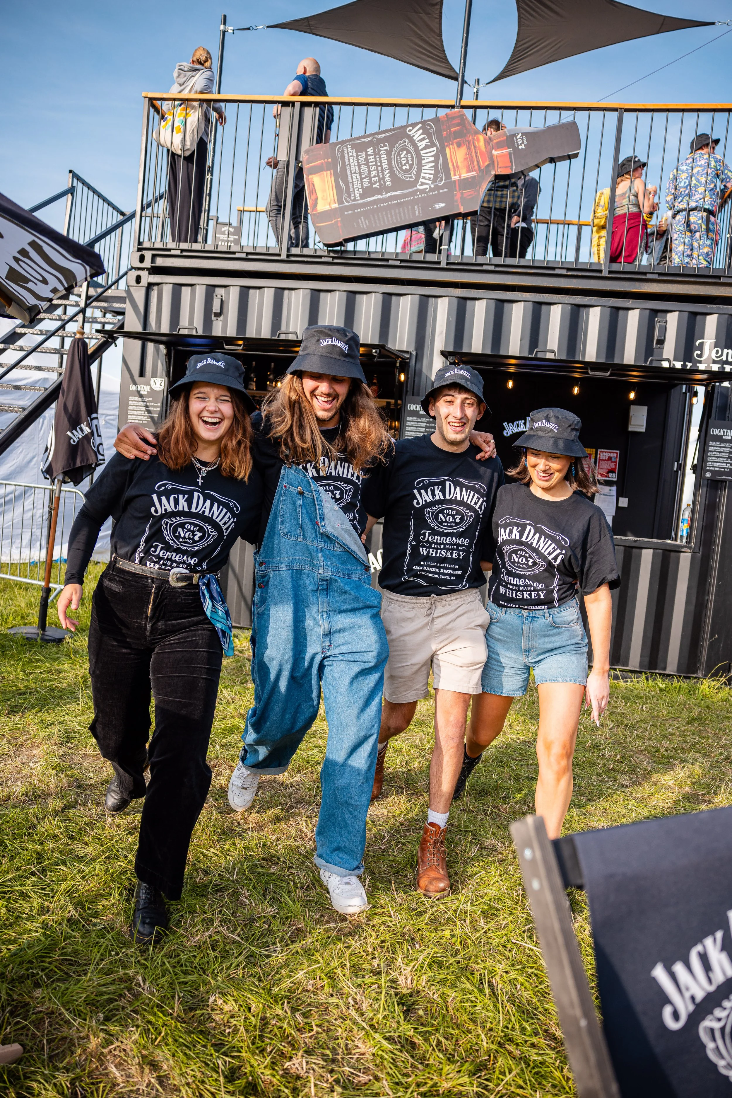 Four friends wearing Jack Daniel's shirts and hats walking on grass, smiling and enjoying an outdoor event with a two-story food and drink stand in the background.