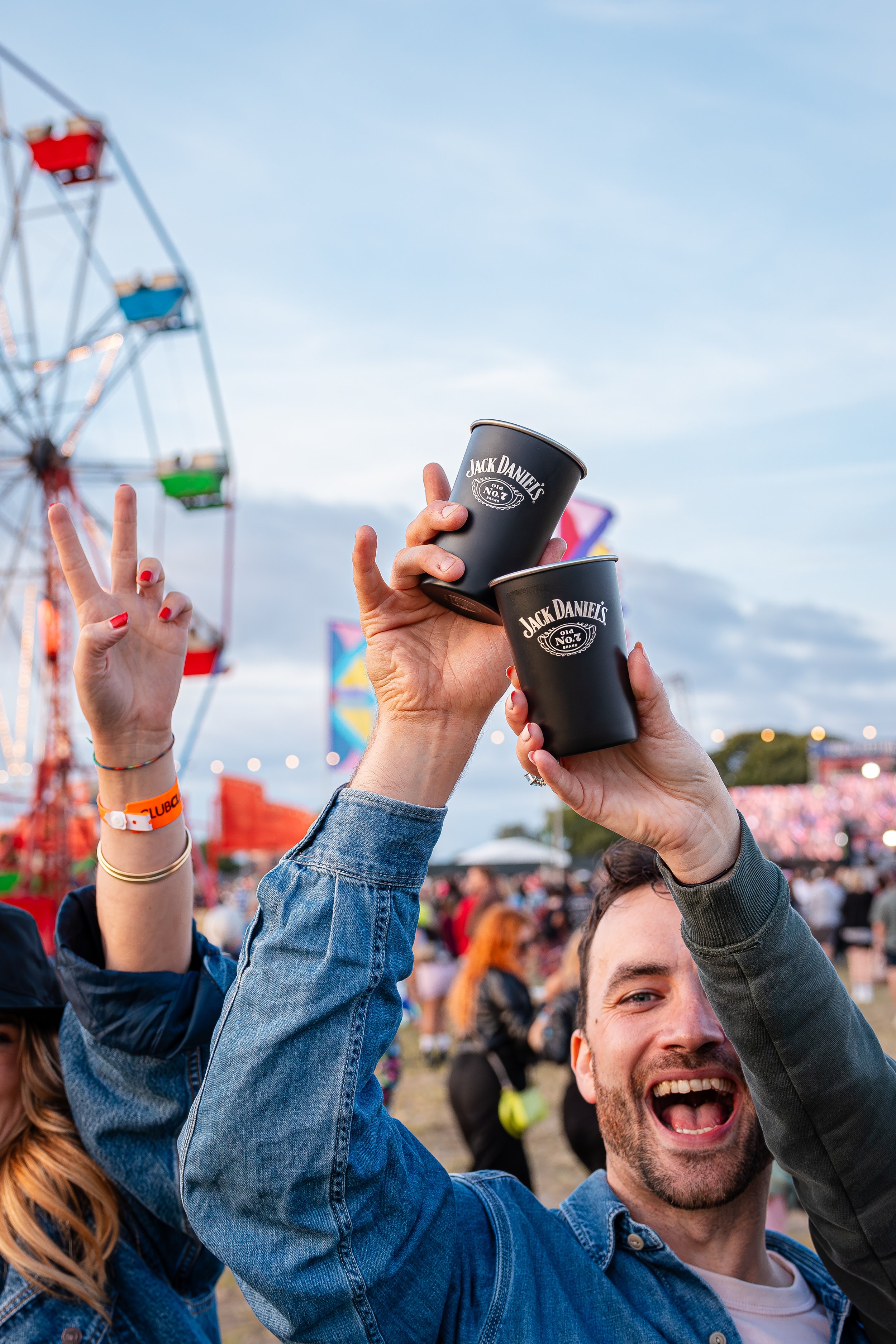 People enjoying a festival or fair, holding up Jack Daniel's branded cups, with a Ferris wheel and crowd in the background.