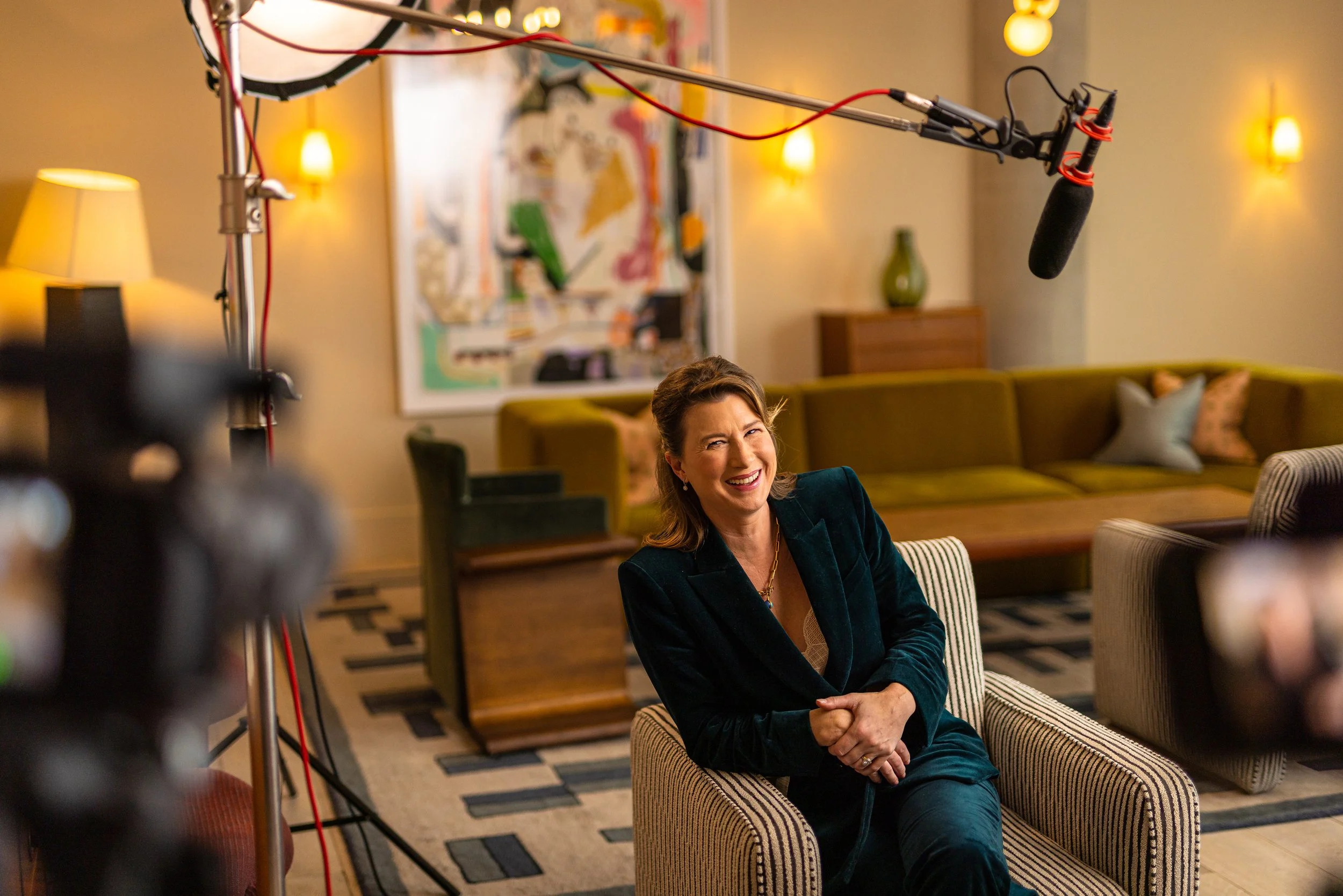 A woman in a dark blazer smiling while sitting on a striped armchair in a warmly lit living room with green and beige sofas, wooden furniture, and abstract artwork on the wall.