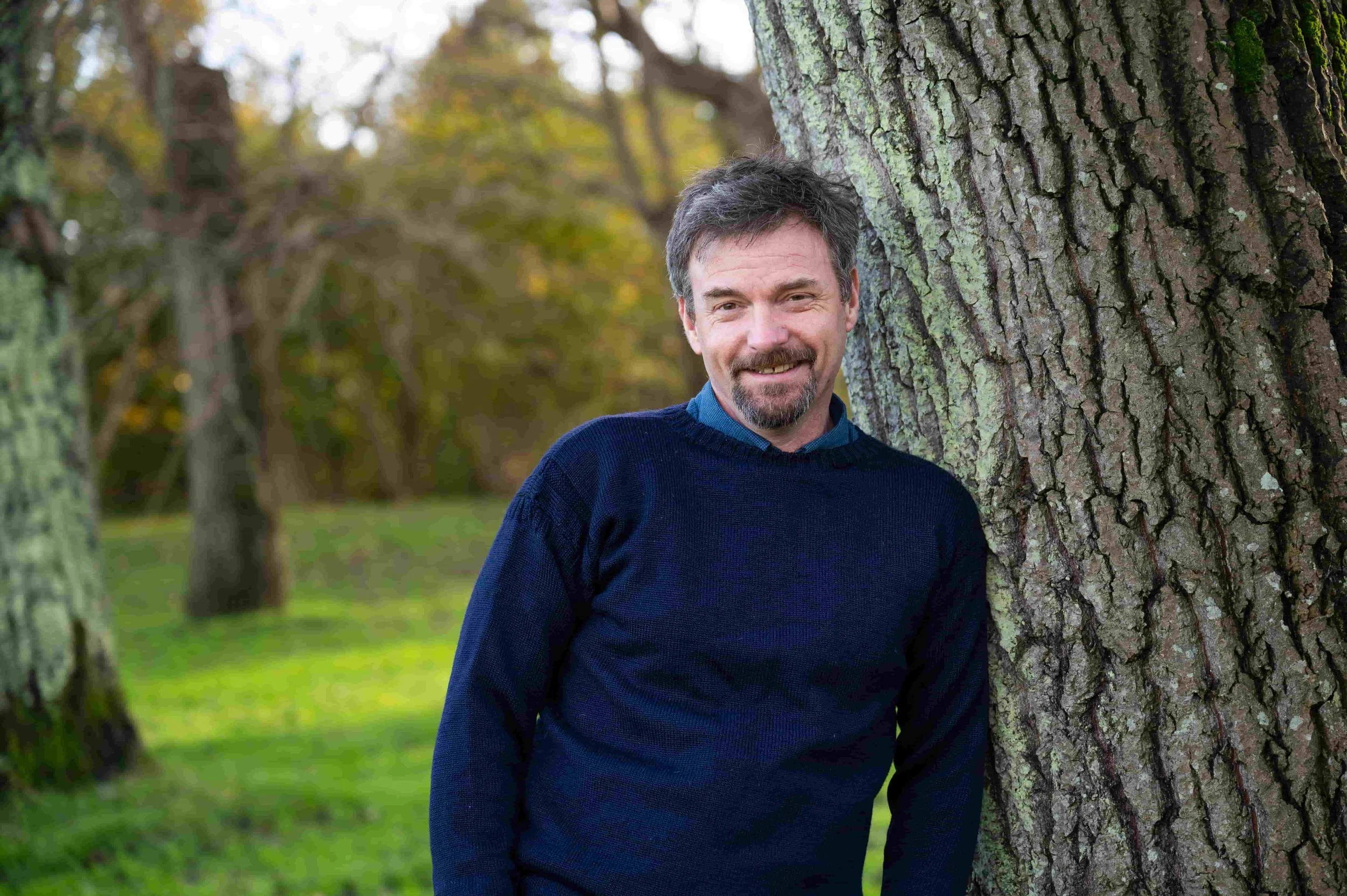 Tony Coward, ADHD Business Coach, smiling while leaning against a large tree in a park during autumn.