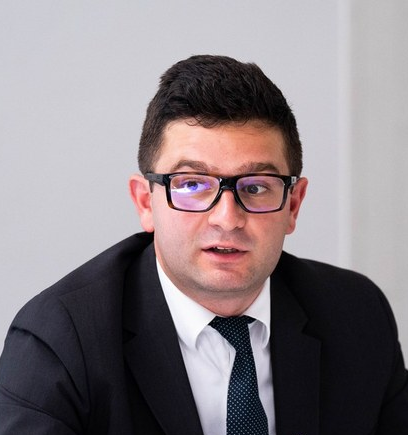 Man in a suit and tie wearing glasses, speaking or listening during a meeting in an office setting.
