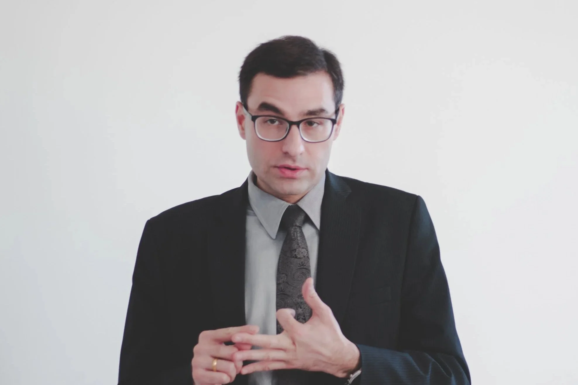 Man wearing glasses in a business suit, gesturing with his hands, against a plain white background.