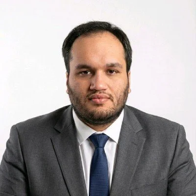 A man with dark hair and a beard fake smiling in front of a plain light-colored wall with ceiling lights, wearing a dark blazer and light-colored shirt.
