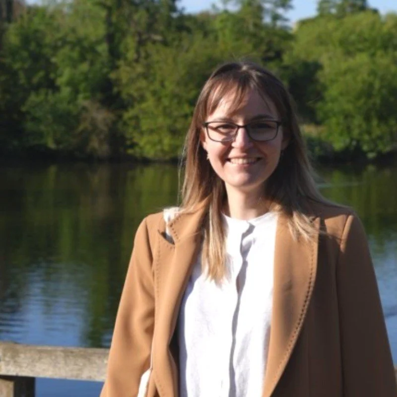 A woman with long brown hair and glasses smiling outdoors near a body of water with trees in the background.