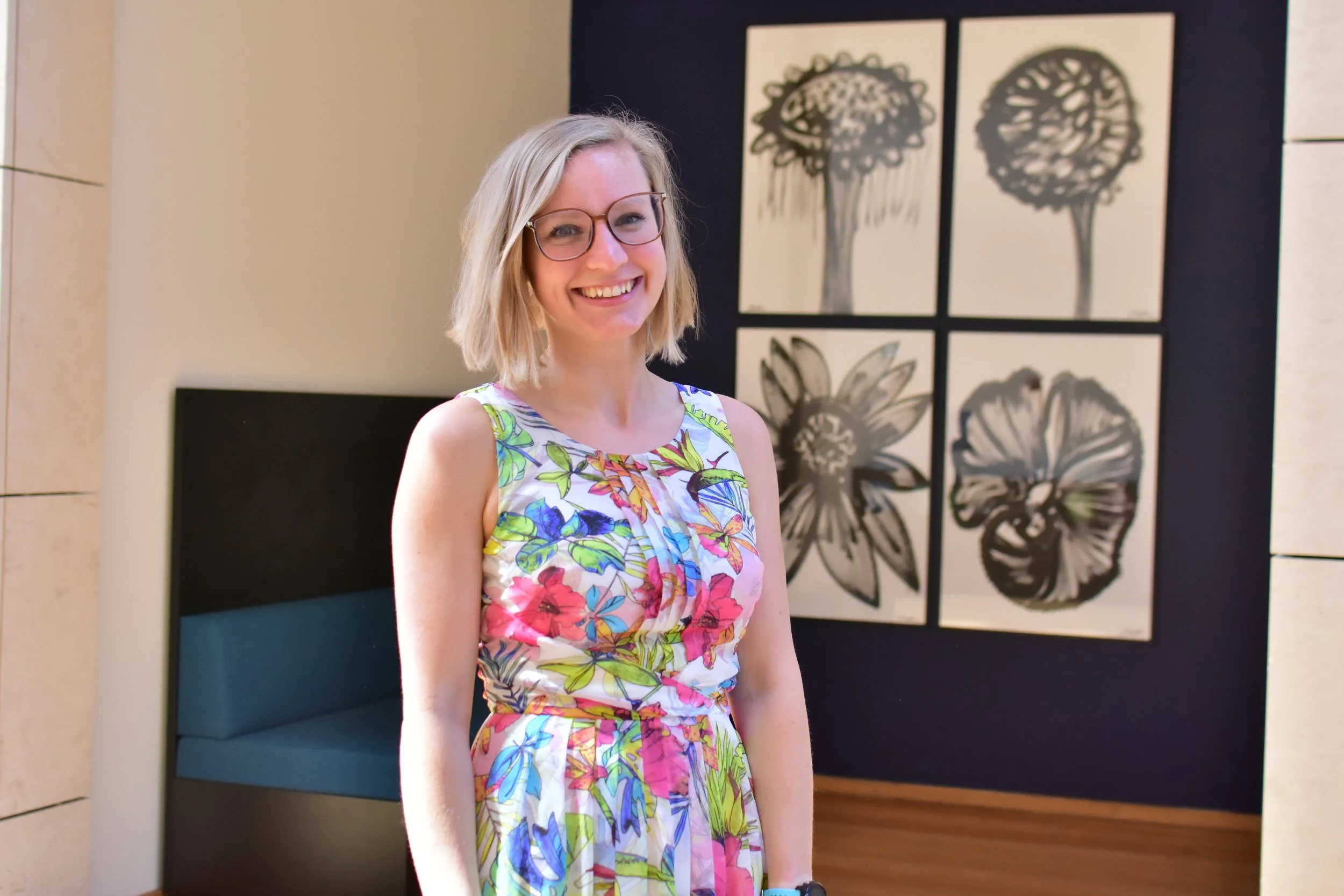 A woman with blonde hair and glasses wearing a colorful floral dress, smiling indoors in front of wall art with black and white floral designs.
