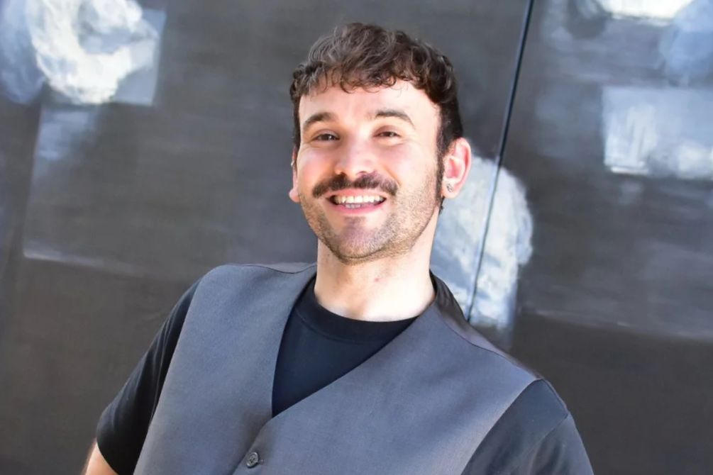 A young man with dark hair and a mustache, smiling, wearing a black T-shirt and a gray vest, standing in front of a black wall with abstract white clouds or paint splashes.