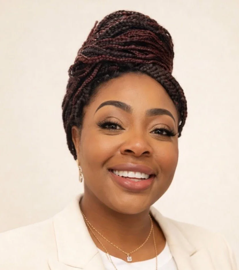Portrait of a smiling woman with braided hair styled in an updo, wearing a white blazer, earrings, and a necklace against a plain background.