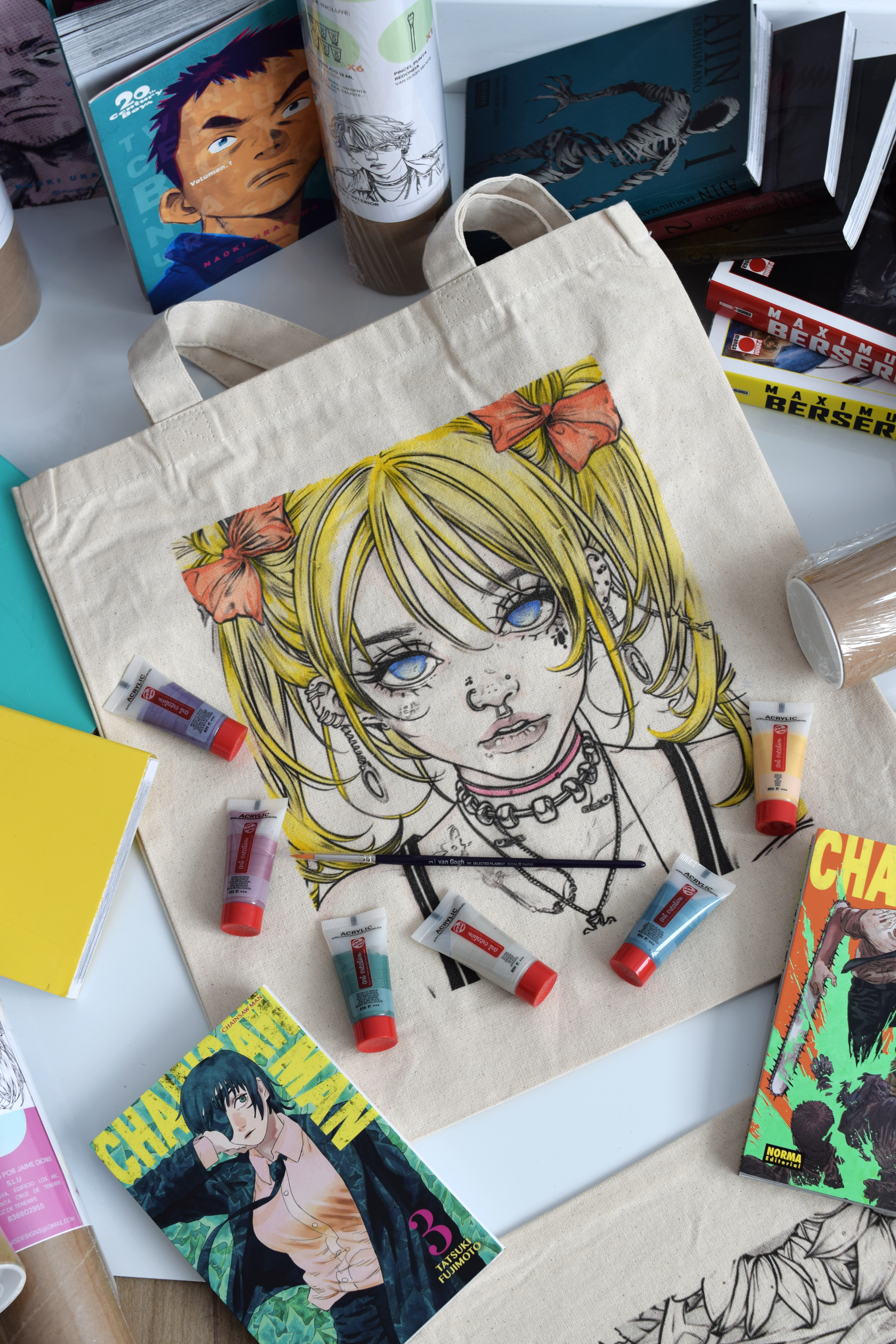 Totebga con diseño impreso pintada con colores arílicos, Ilustración de una joven con cabello rubio, ojos azules, accesorios y maquillaje, rodeada de tubos de pintura y libros de cómics en una mesa de arte.