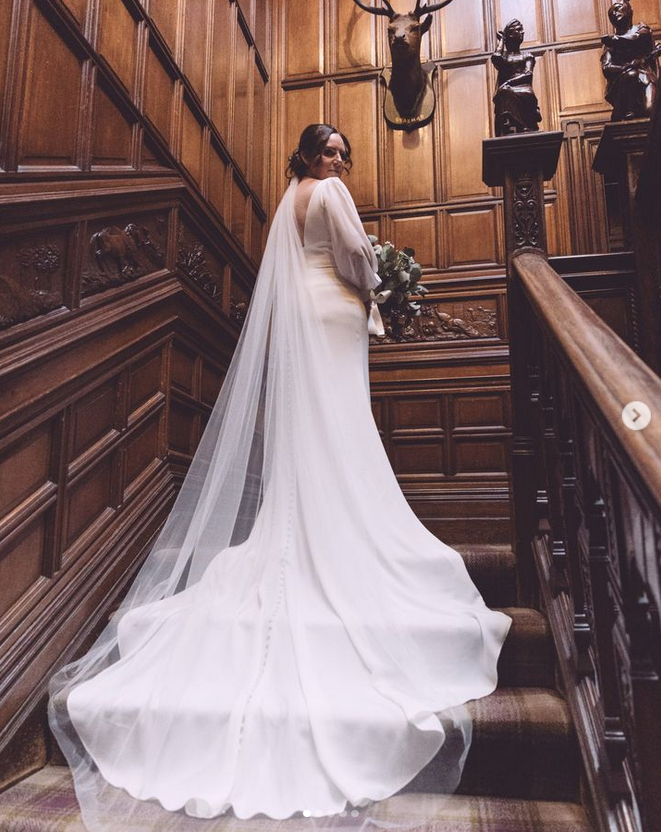 Bride standing on a wooden staircase in a grand, wood-paneled room with carved wood details and statues. She is wearing a white wedding gown with a long train and veil, holding a bouquet and looking over her shoulder.
