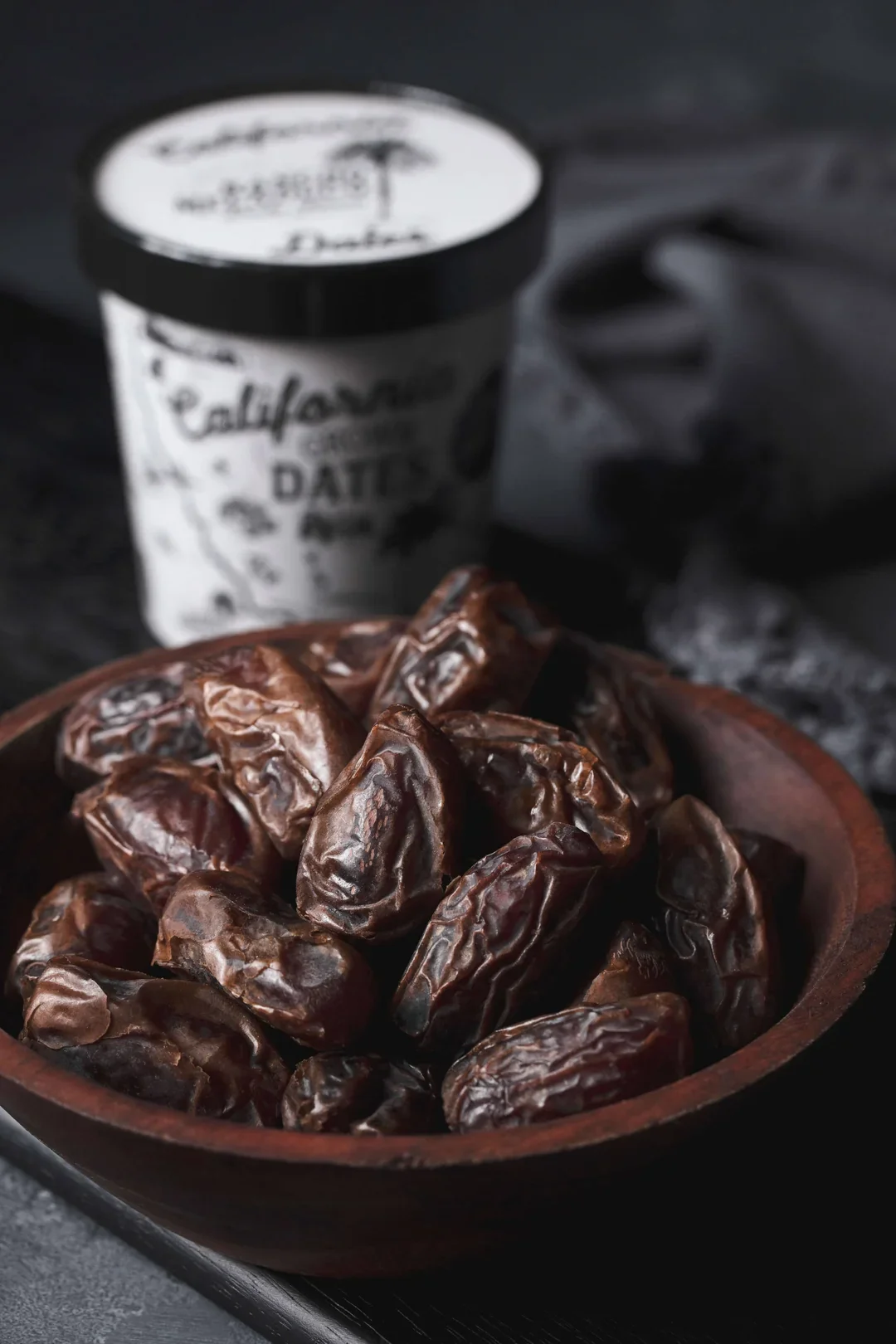 A wooden bowl filled with wrinkled, dark brown dates in the foreground. In the background, there is a blurry paper cup with a black lid, likely containing a beverage, resting on a dark surface.