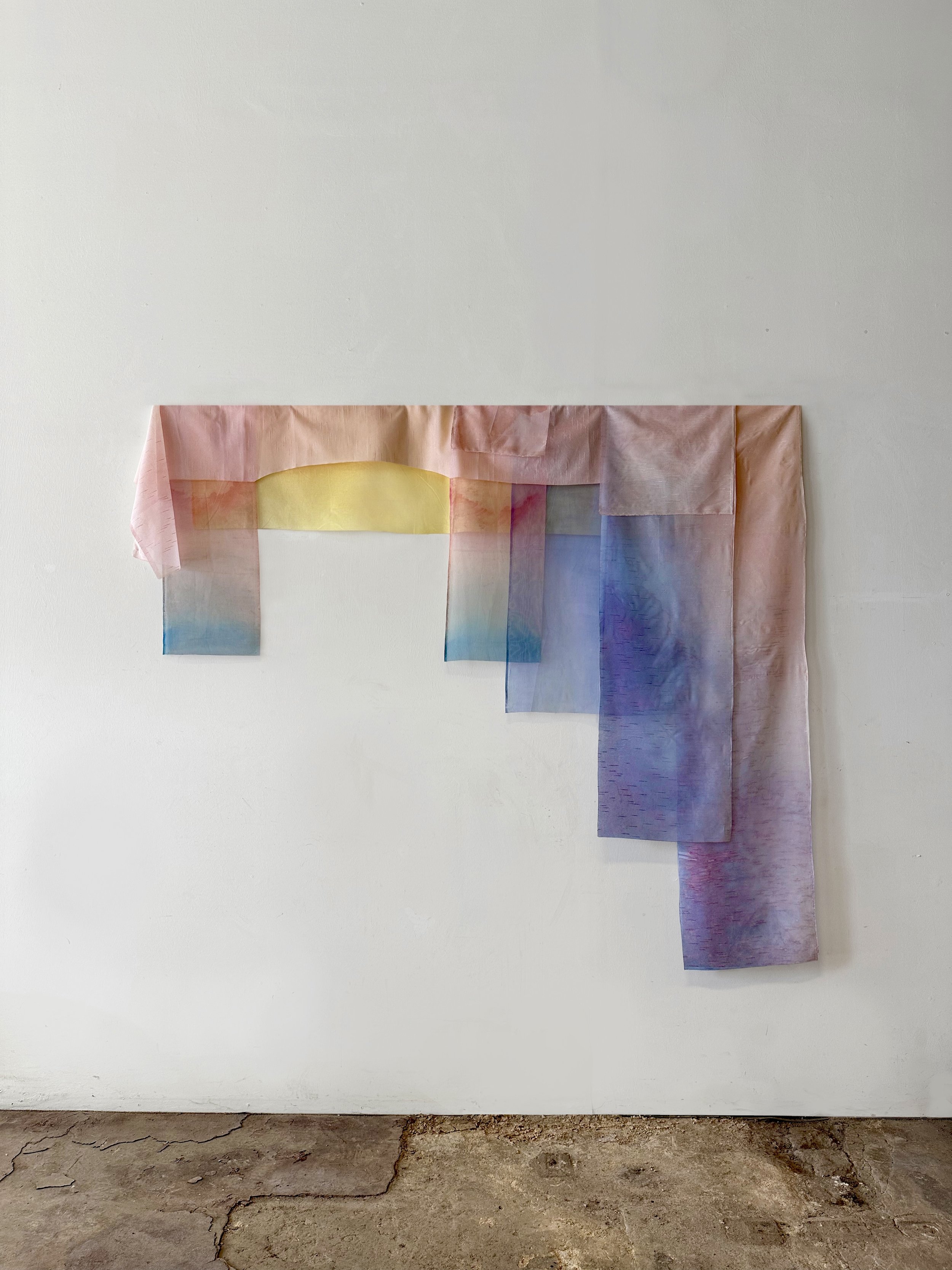 Colorful, semi-transparent fabric strips hanging on a wall, with a white wall background and a cracked concrete floor at the bottom.