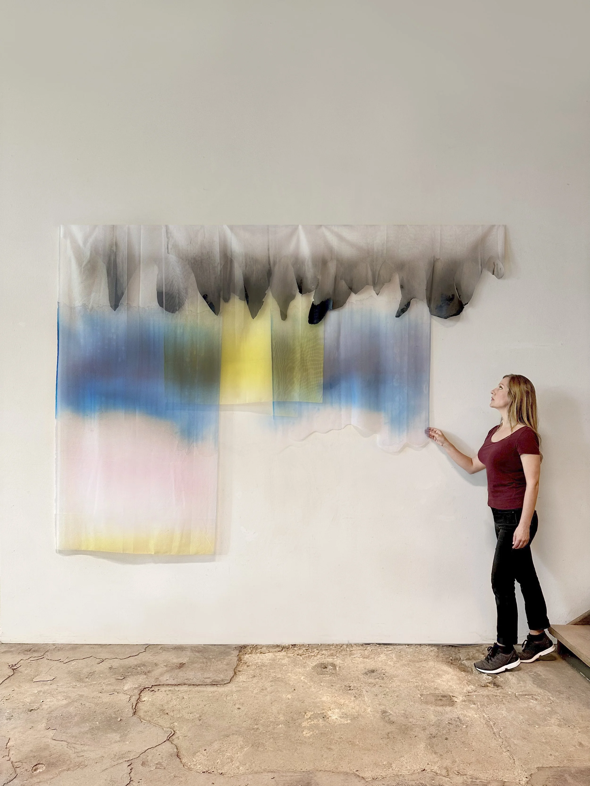 A woman with long blonde hair wearing a maroon shirt, black pants, and athletic shoes standing next to a large abstract watercolor painting on a white wall. The painting features soft blending colors, including blue, green, yellow, and pink, with a t