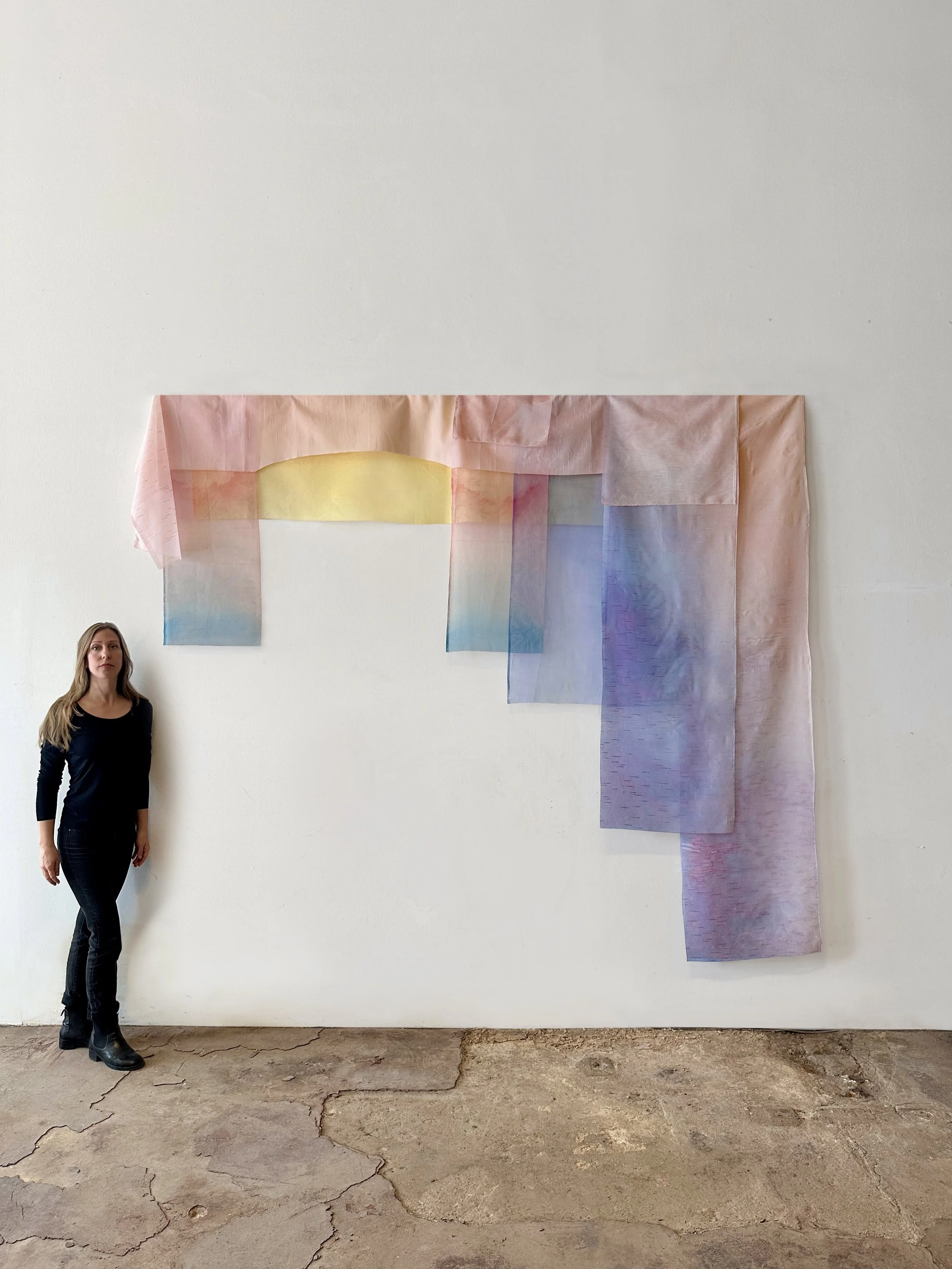 A woman standing next to a large wall hanging made of pastel-colored, translucent fabric pieces arranged in a rectangular formation.