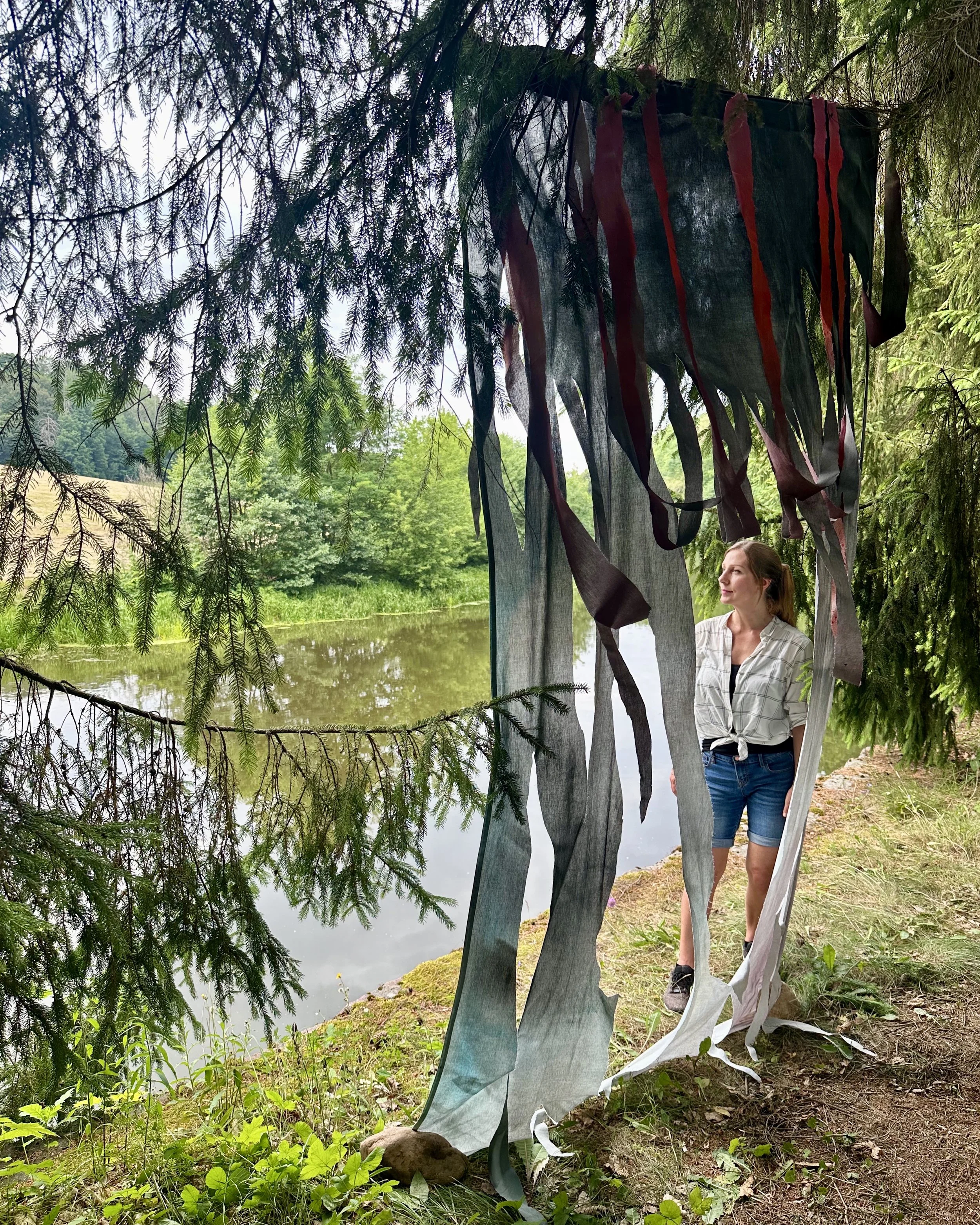 A woman standing outdoors near a body of water, surrounded by trees and greenery, partially obscured by large, dark fabric strips hanging from a tree with red accents.