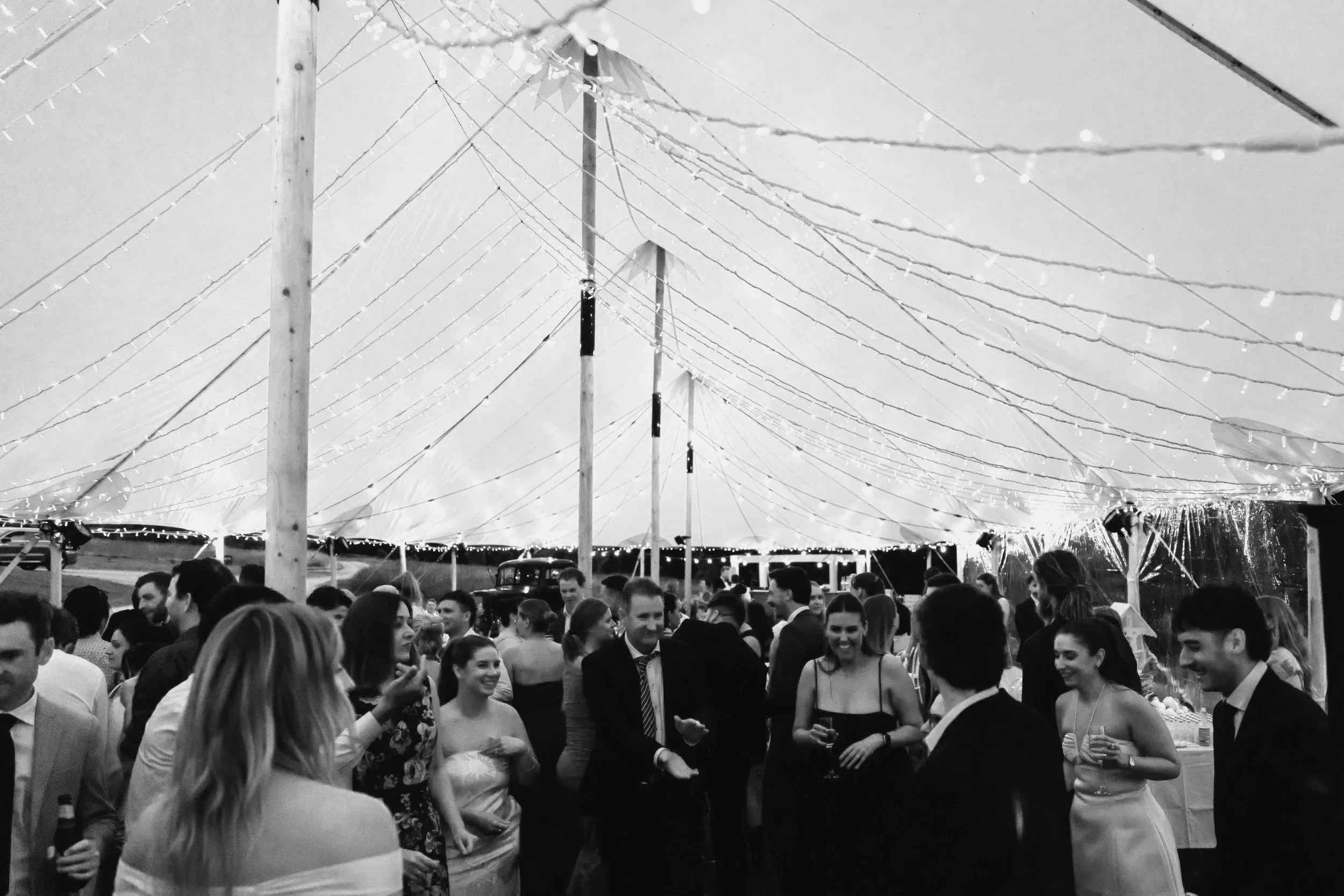 Wedding dance floor, group of friends dancing in a circle laughing. Big marquee in background with strings of fairy lights.