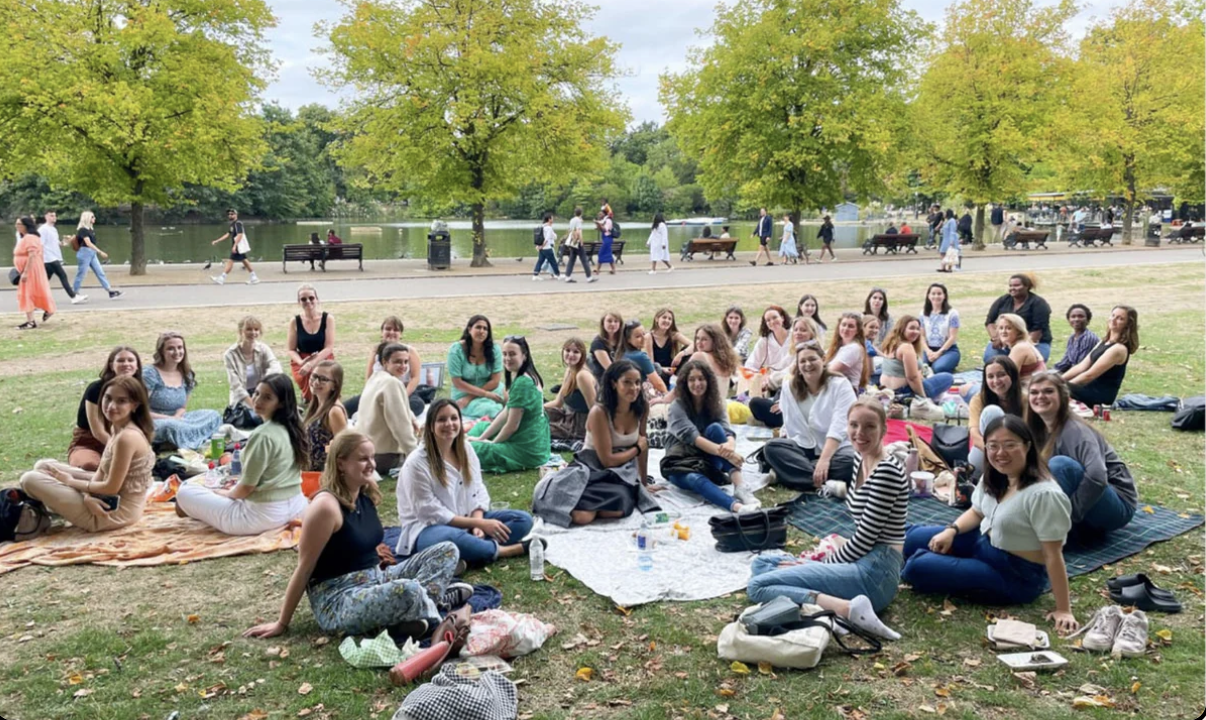 A large group of people sitting on the grass and enjoying a picnic in a park. Some are seated on blankets, and others are walking along a paved path behind them. There are trees and a lake in the background.