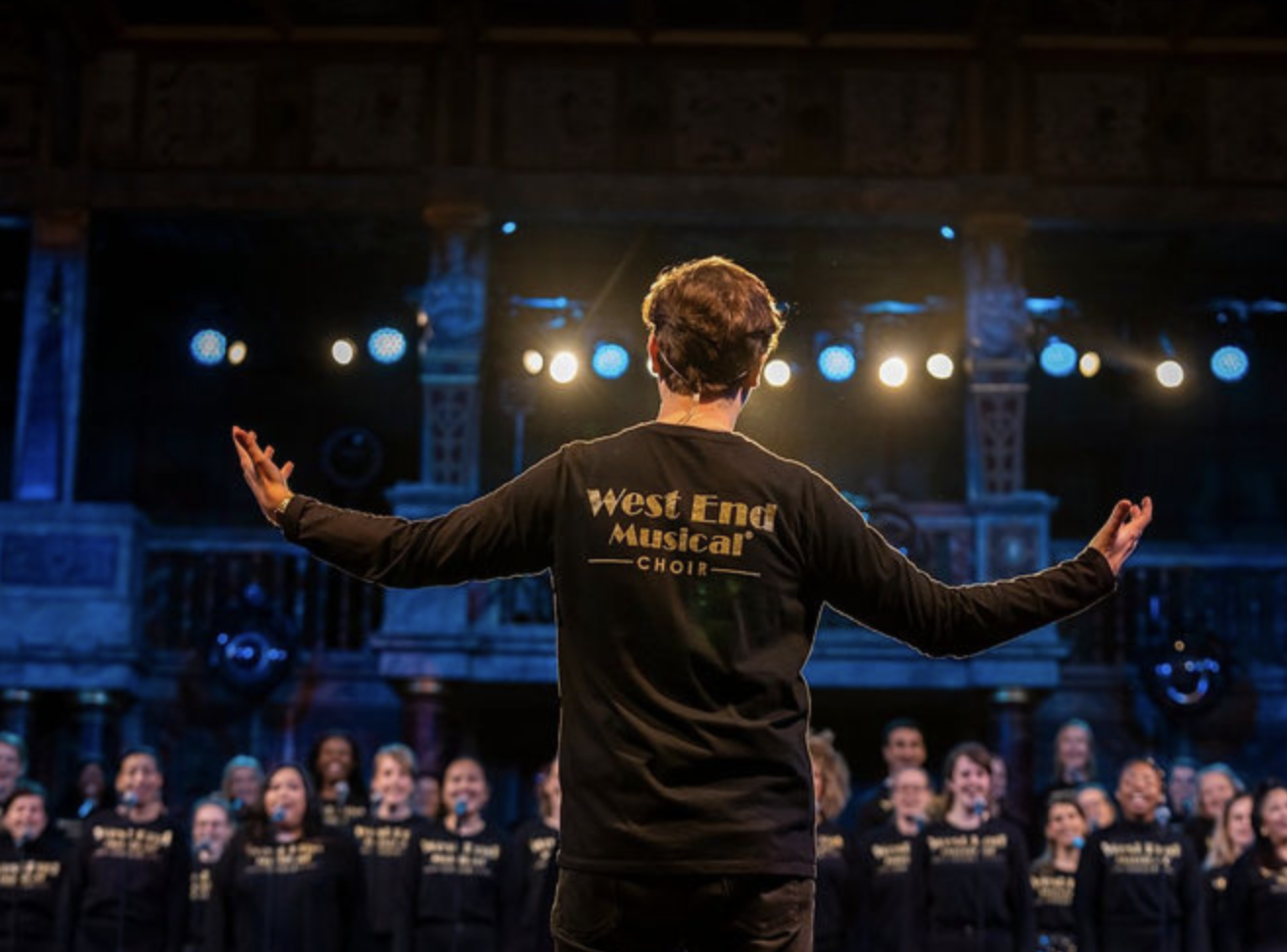 A choir conductor standing with arms outstretched, facing a choir of singers in a theater or concert hall, wearing a black jacket with 'West End Musical Choir' printed on the back, with stage lights and architectural details in the background.