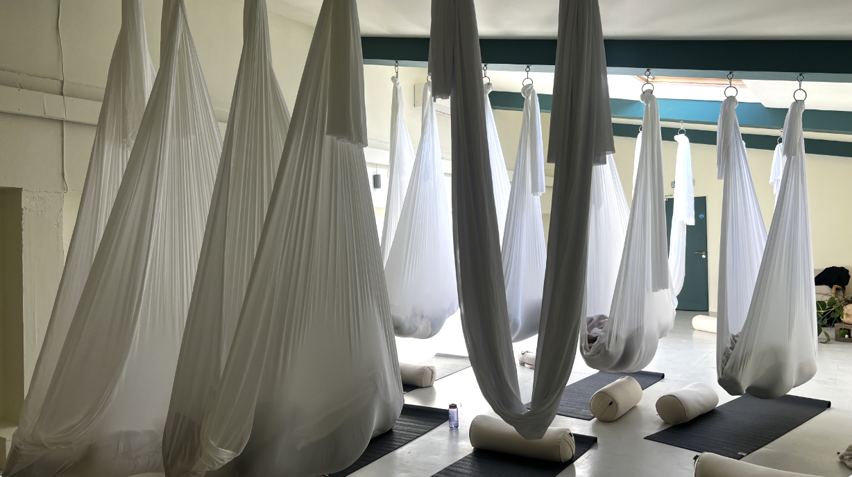 A room with multiple white fabric hanging yoga hammocks suspended from the ceiling, with yoga mats and bolsters on the floor.