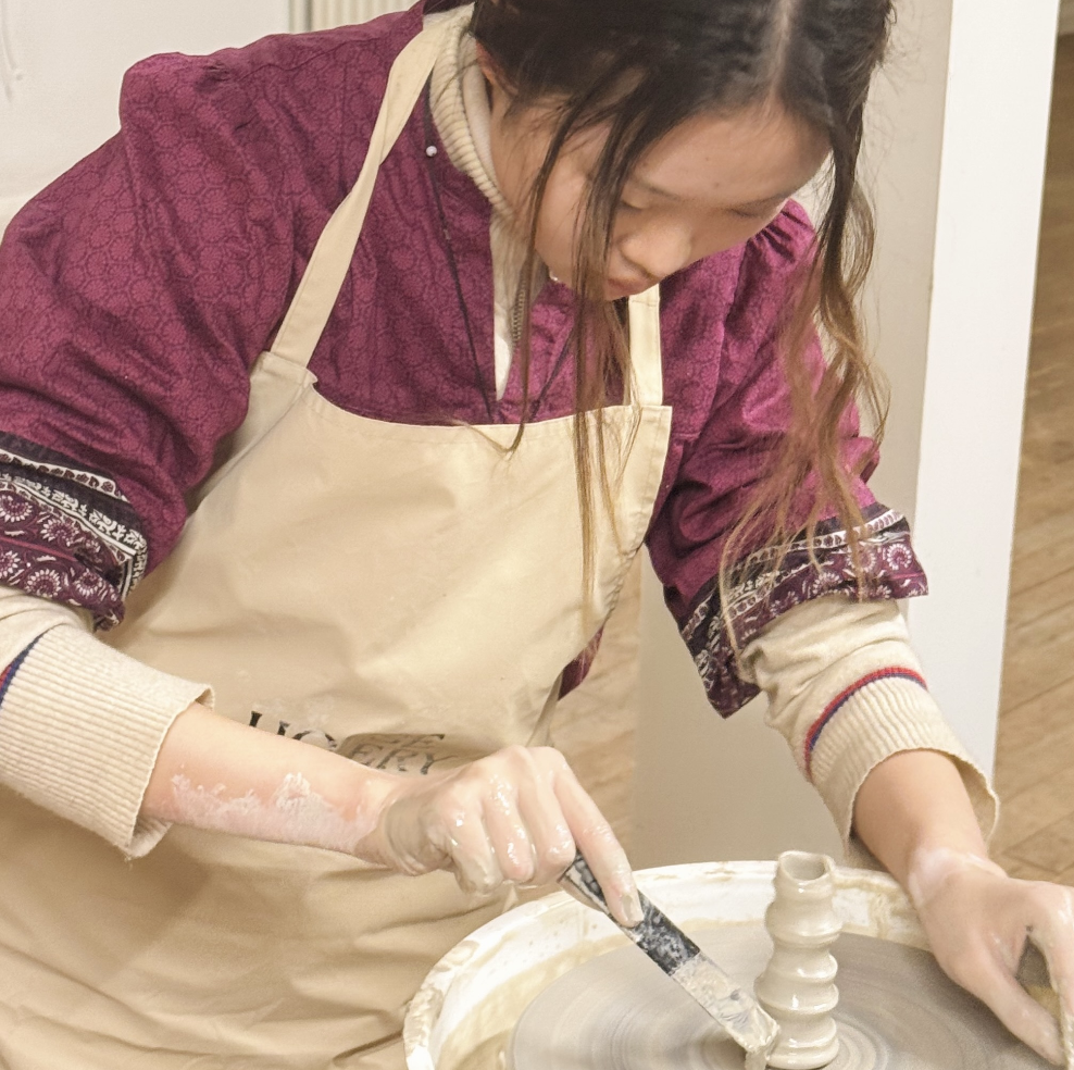 A woman wearing a beige apron and a patterned purple jacket is using a pottery wheel to shape clay into a vessel.