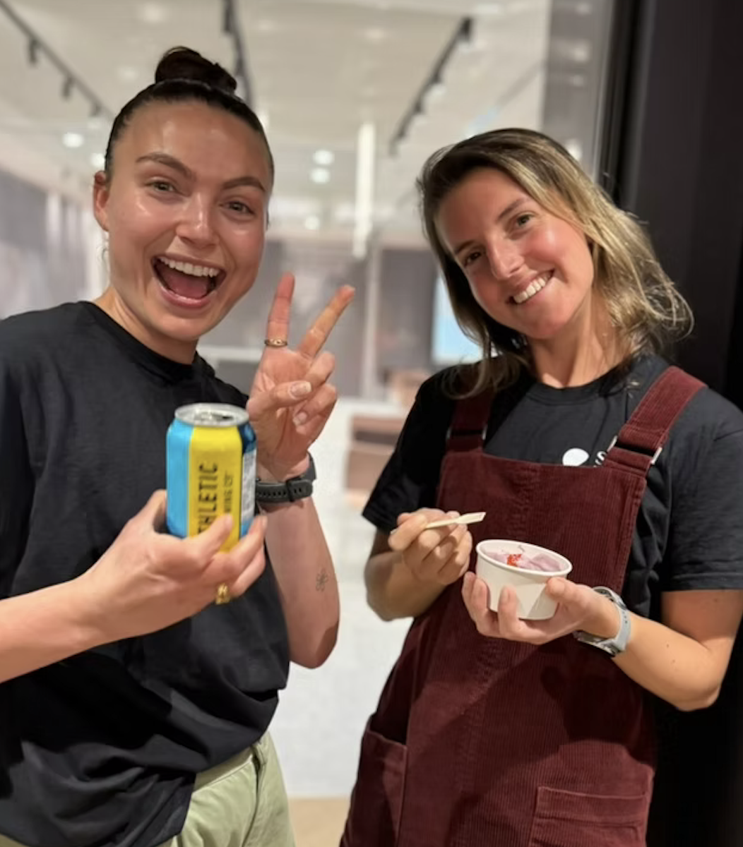 Two women smiling for a photo, one holding a can of sparkling water and the other eating ice cream in a small cup, indoors with a modern background.