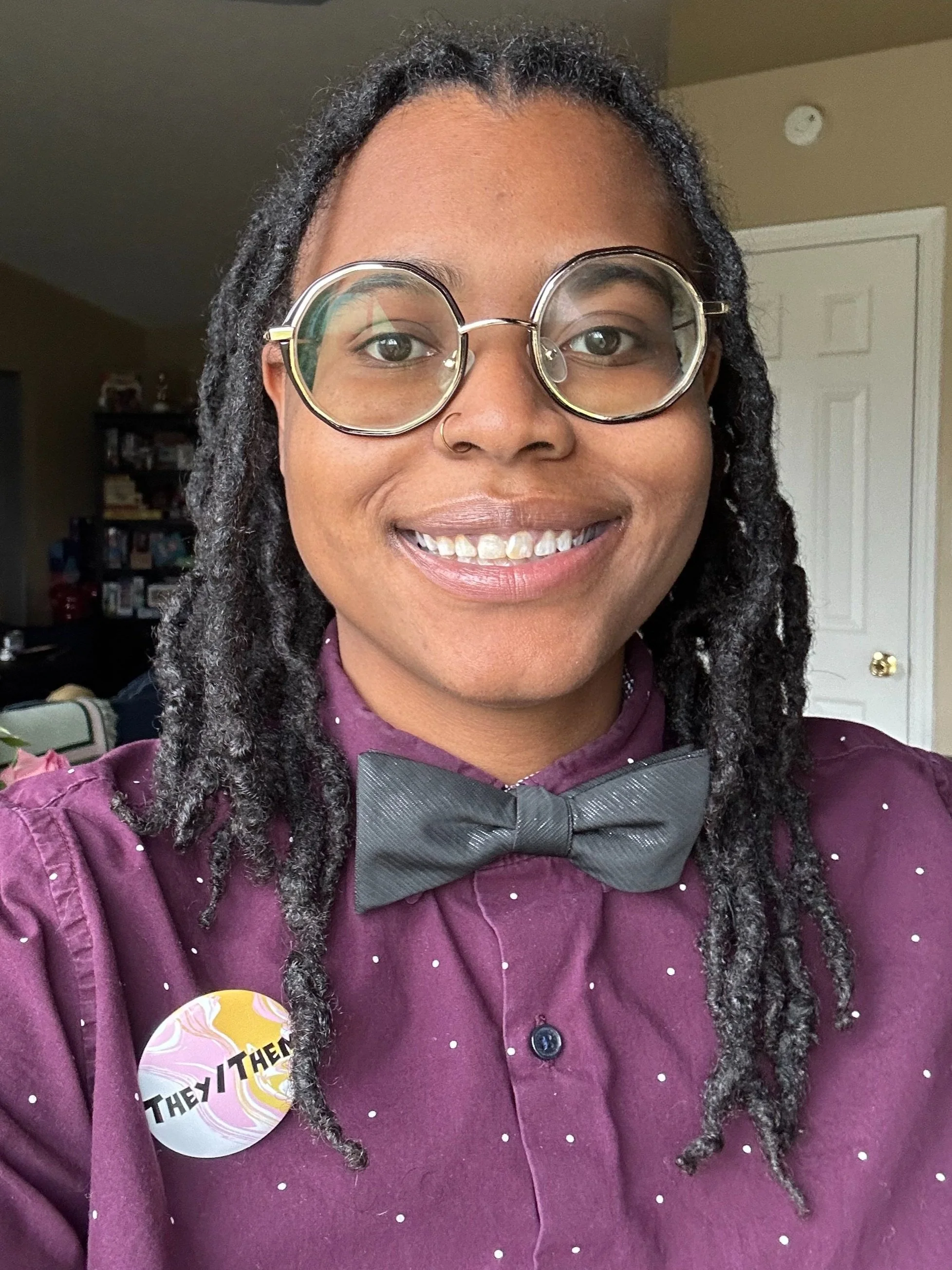 A person with glasses, a nose ring, and dreadlocks wearing a maroon shirt with white polka dots and a black bowtie, smiling at the camera.