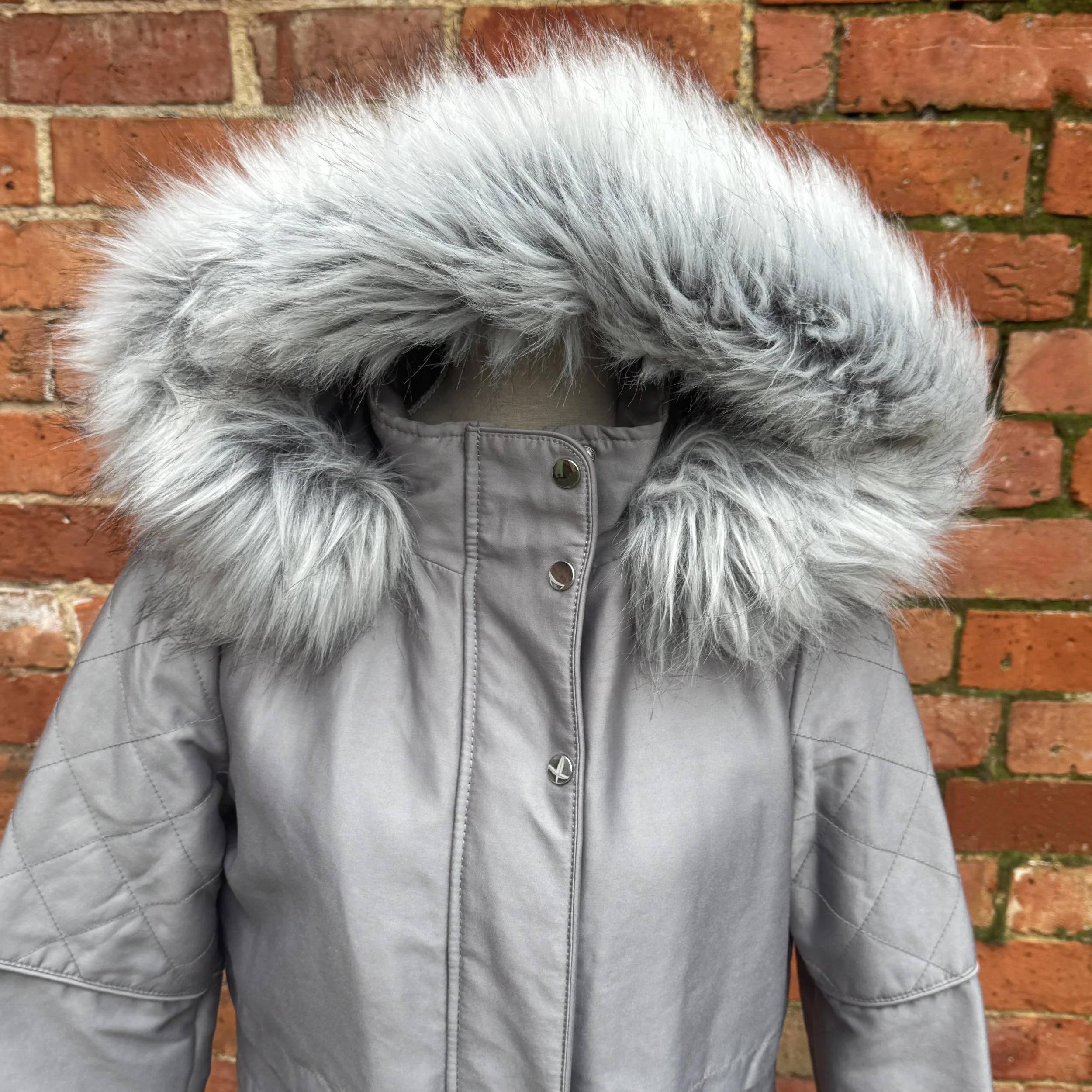 A light gray winter coat with a large, fluffy gray and white faux fur-lined hood against a red brick wall.