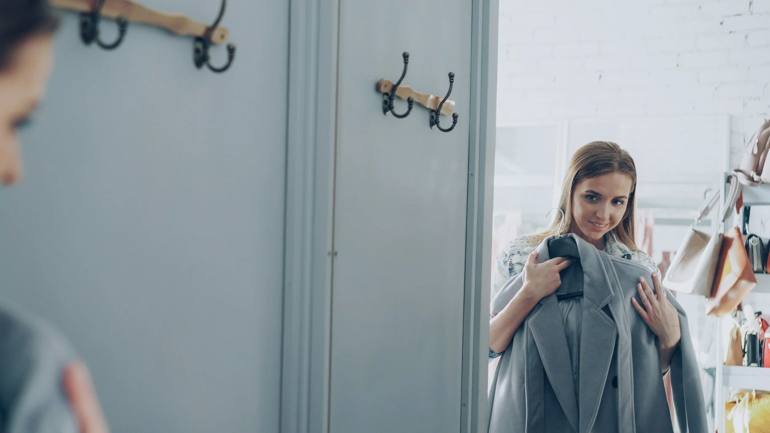 A woman shopping for a gray jacket in a store's dressing room, holding the jacket and smiling.