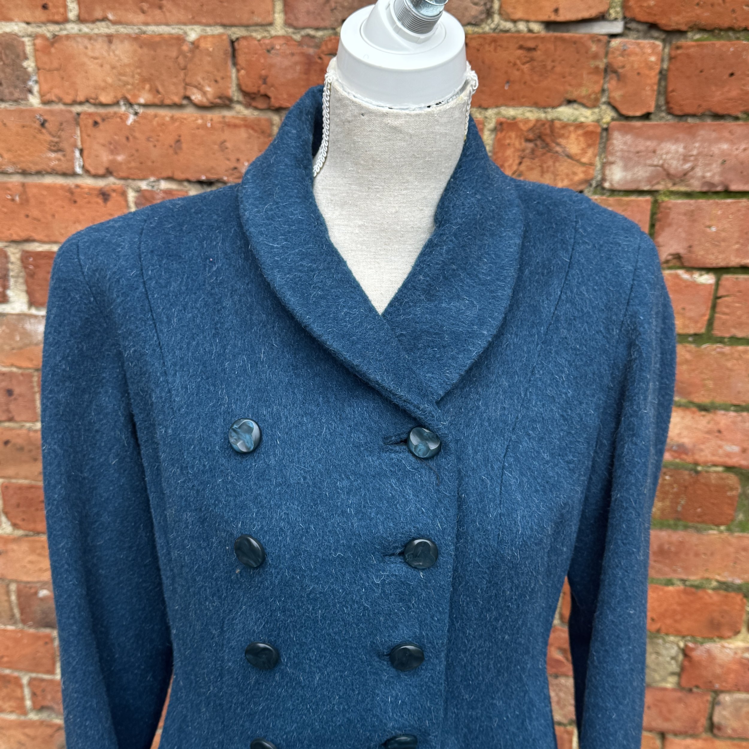 Blue double-breasted wool coat with large buttons displayed on a mannequin against a brick wall background.