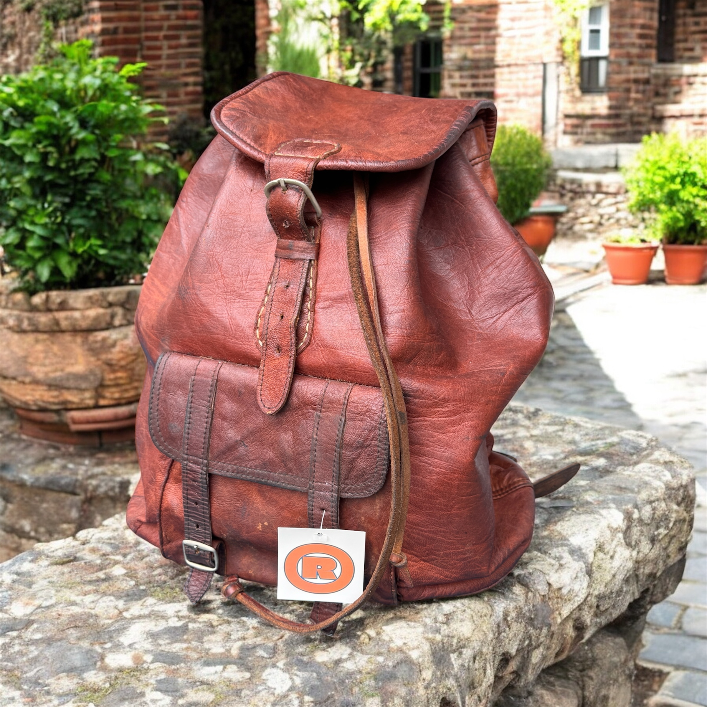 An old, worn leather backpack resting on a stone surface outdoors, with a plant and brick building in the background.