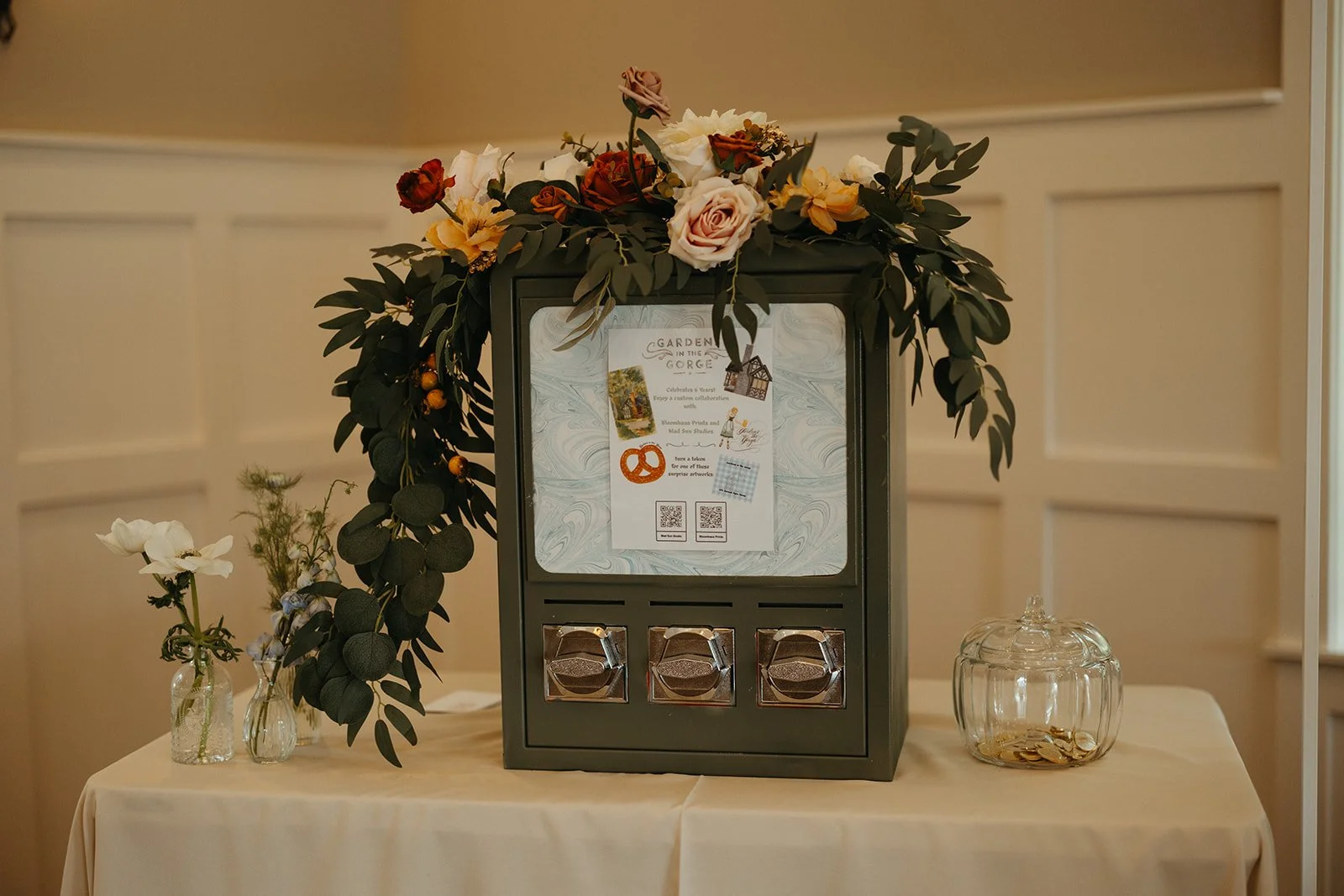 A donation box decorated with a floral arrangement on top, placed on a table with small vases and a jar of coins inside.