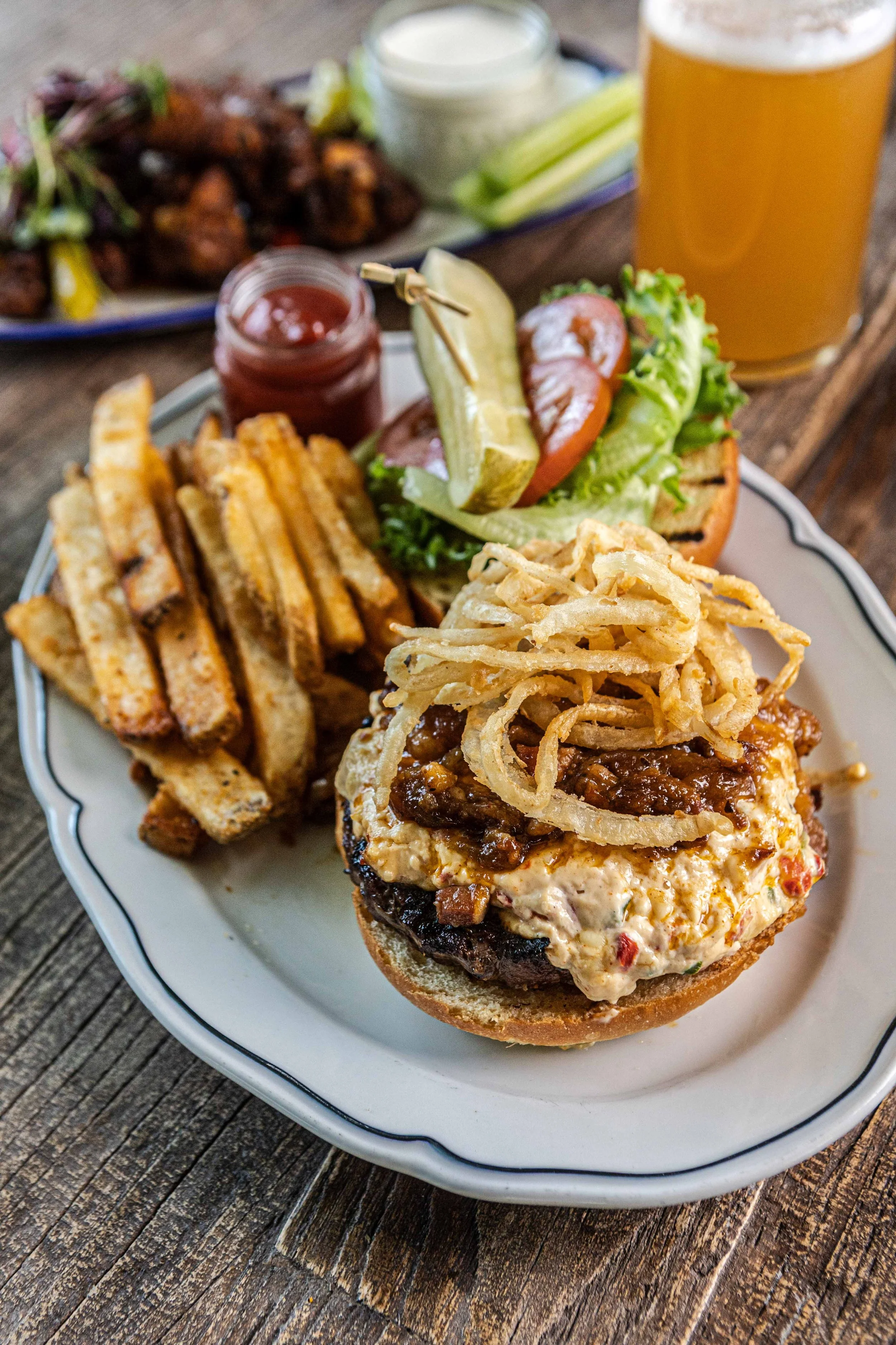 Close-up of a plate with a gourmet cheeseburger topped with fried onion strings, alongside crispy French fries, and a miniature sandwich with lettuce, tomato, and pickle. In the background, there are additional dishes, a jar of barbecue sauce, and a glass of beer on a rustic wooden table.