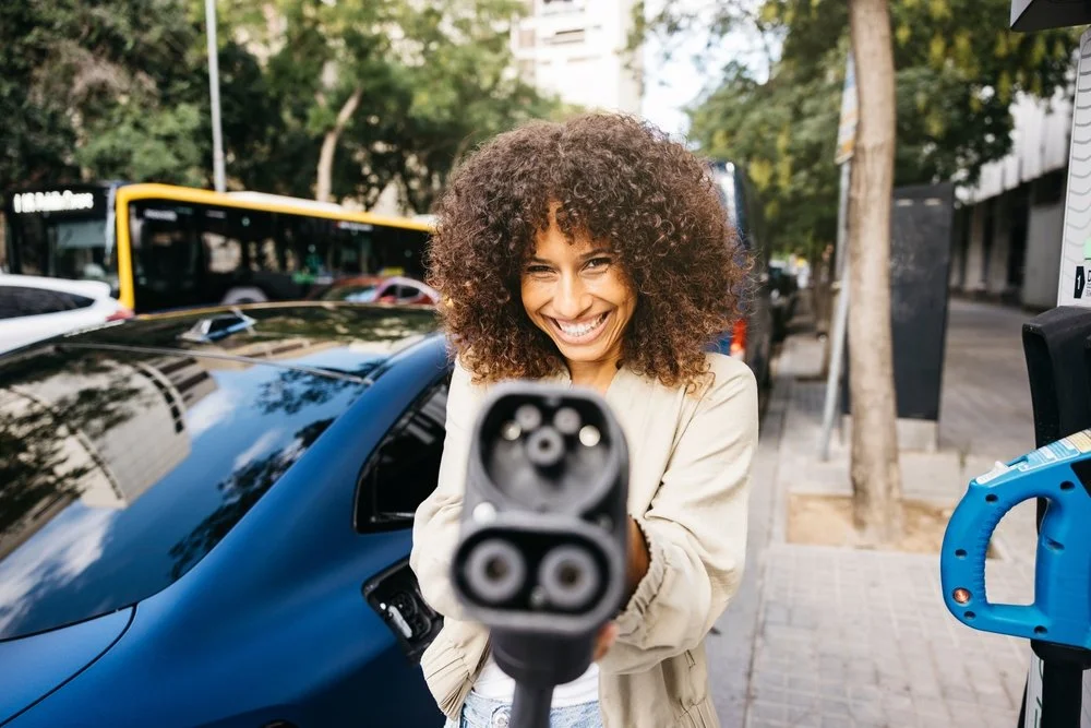 A smiling woman holding out a Type 2 EV charging cable towards the camera next to a blue electric car on a city street.