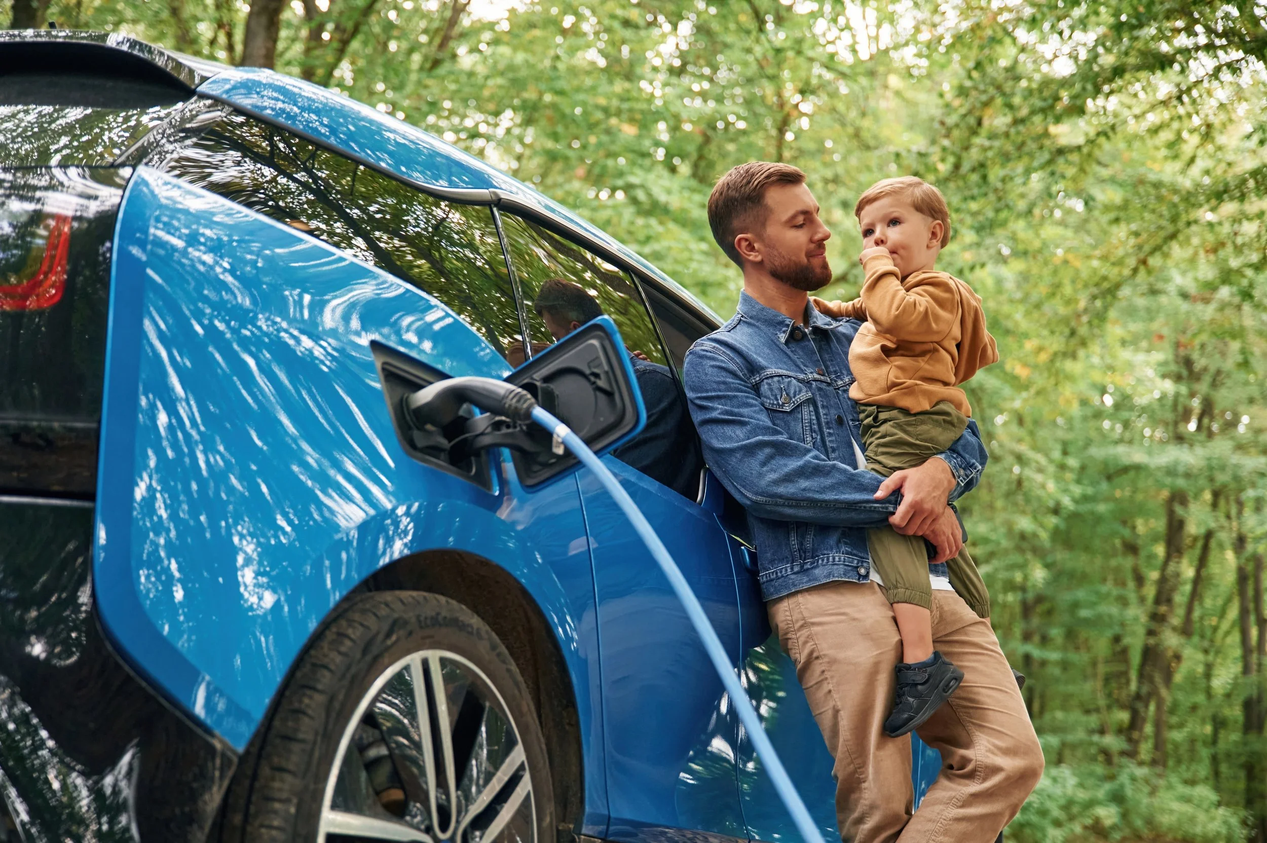 Close-up of a blue electric vehicle (EV) charging in a wooded park area with a man and toddler standing nearby.