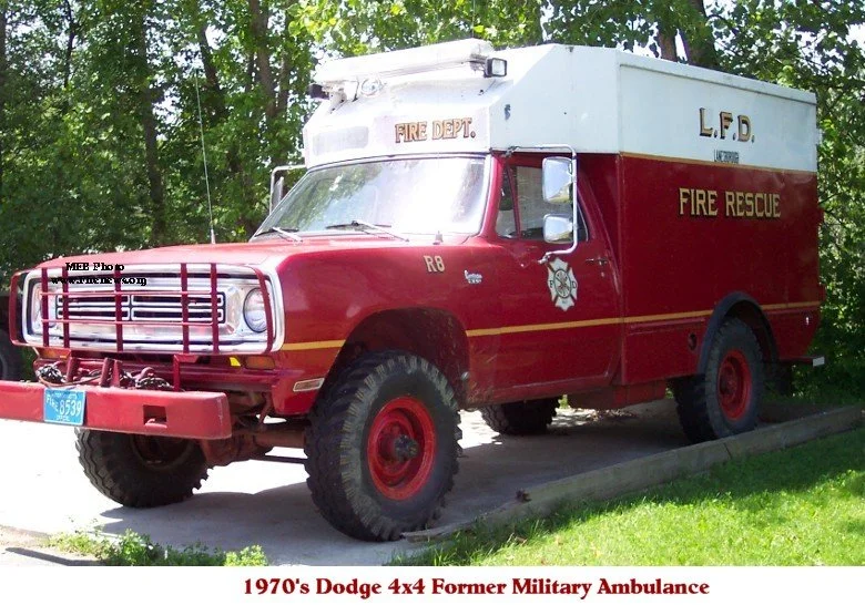 A 1970s Dodge 4x4 fire department military ambulance vehicle, painted red and white, with fire rescue markings, large tires, and emergency lights, parked outdoors on a slight incline with trees in the background.