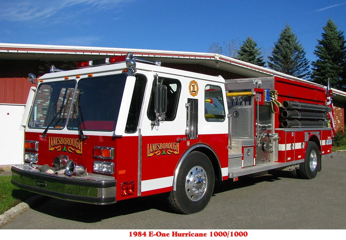 Red and white fire truck with equipment and hoses on the side, parked outside a building with trees in the background.